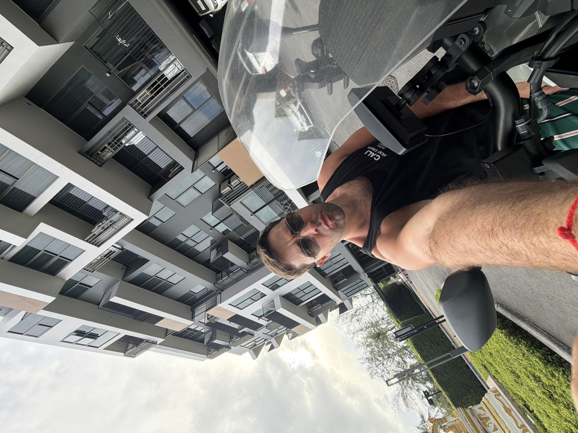 A man wearing sunglasses and a black tank top with the text "CALI WEST COAST SAN DIEGO CA" is seated on a motorcycle. The man is holding the camera up to take a selfie. The background features a multi-story building with windows and a partly cloudy sky. A street with green hedges and what appears to be ornate Thai decorative elements are visible. Based on the elements in the image, the location appears to be Choeng Thale, Thailand.