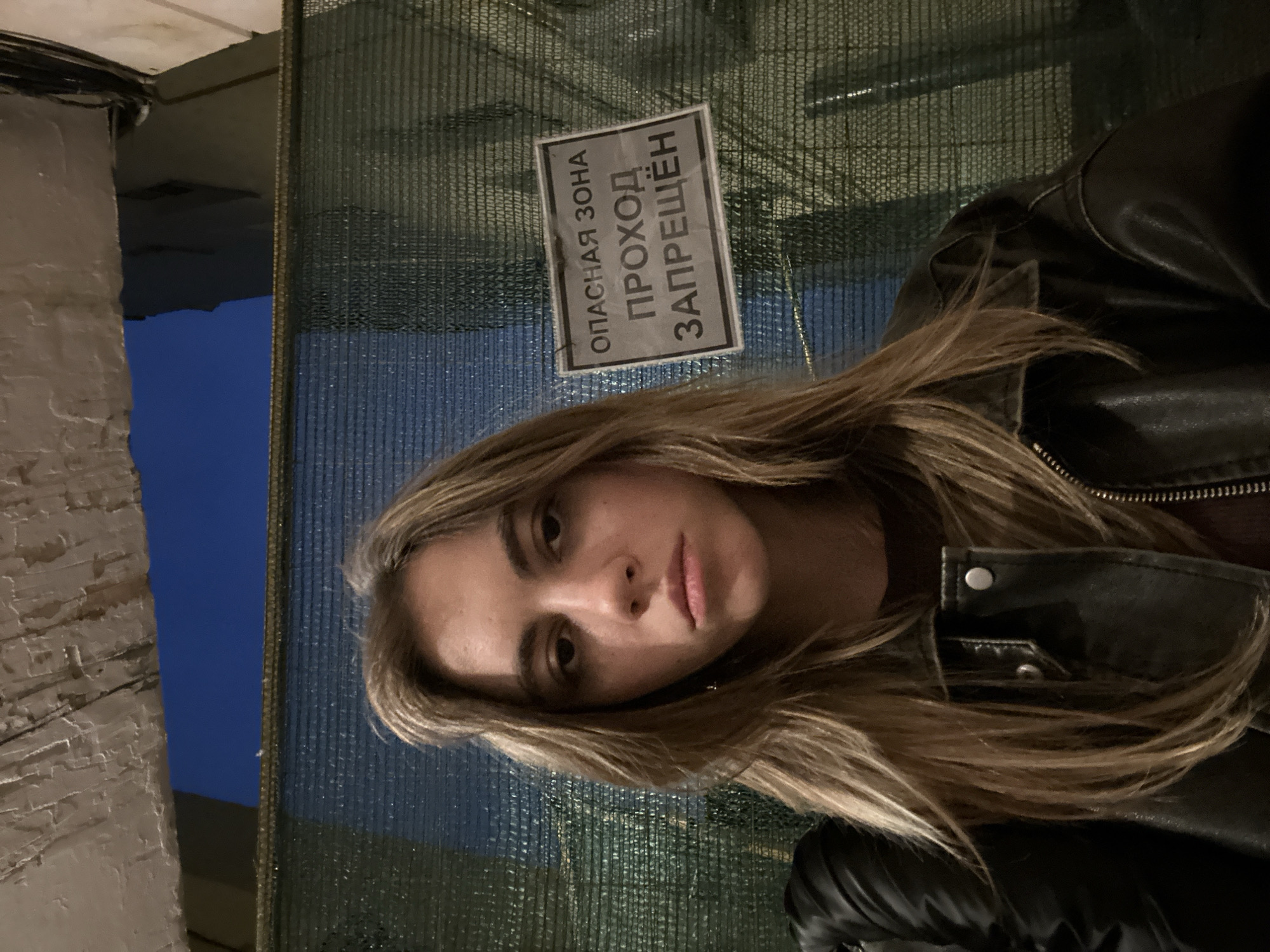 A young woman with long, light brown hair and freckles is pictured from the chest up, looking directly at the camera. She is wearing a dark leather jacket over a dark shirt. Behind her, a green mesh barrier is visible, with a sign on it that reads "ОПАСНАЯ ЗОНА ПРОХОД ЗАПРЕЩЁН" which translates to "DANGER ZONE PASSAGE PROHIBITED". Above the barrier, a glimpse of a dark blue sky and part of a building structure are visible. The lighting suggests it is dusk or night. The context of the sign implies the location is a restricted or construction area within Moscow, Russia.
