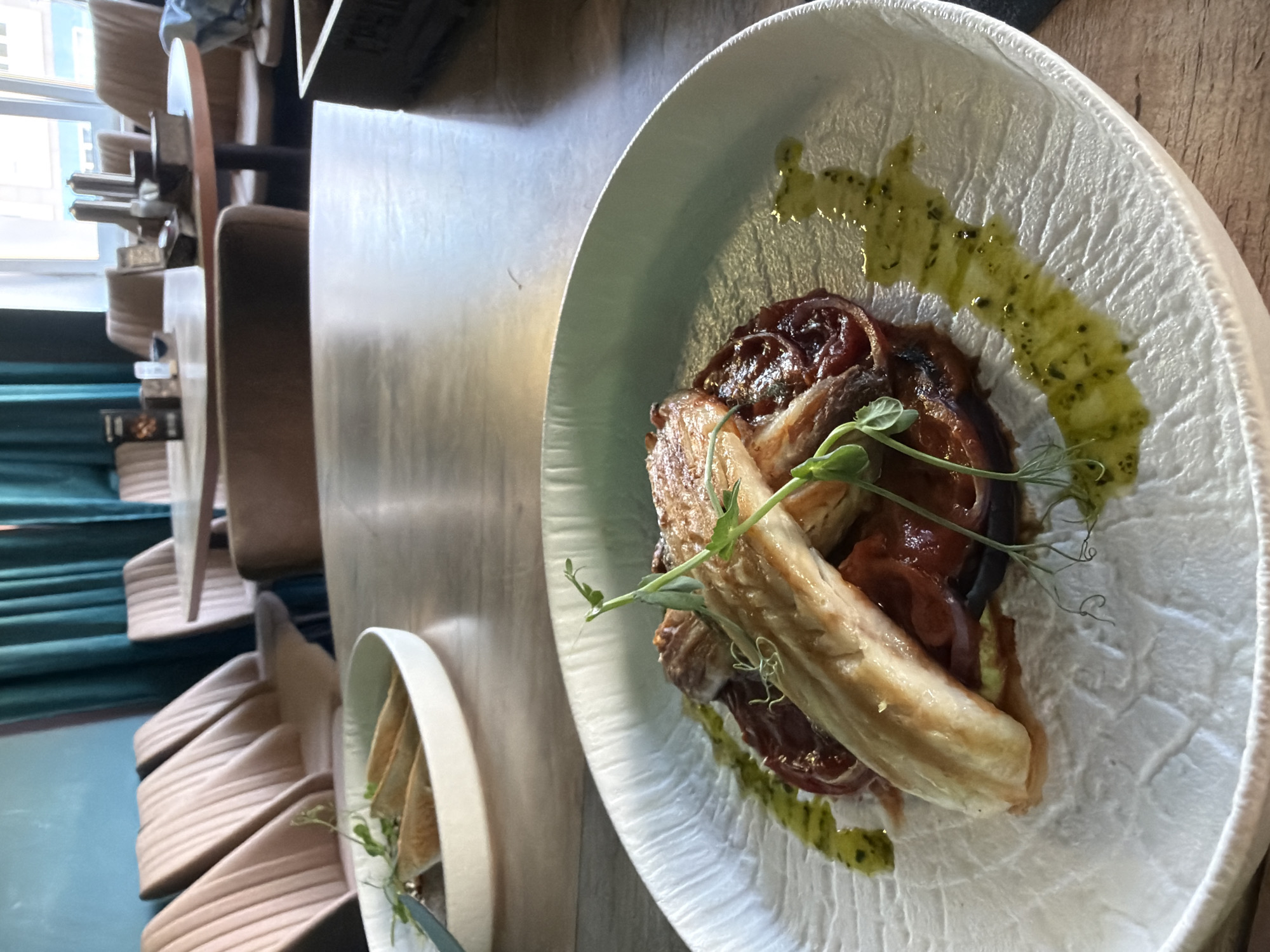 The image depicts a plate of food on a table, possibly in a restaurant setting. The dish includes what appears to be meat, tomato, and other ingredients, garnished with a green sauce and sprouts. The plate has a textured, white surface. Another plate, possibly containing bread, is also visible on the table. The background shows chairs and a table in what looks like a dining establishment. The location in the city of Chaikovsky, Russia, cannot be confirmed from the image alone.