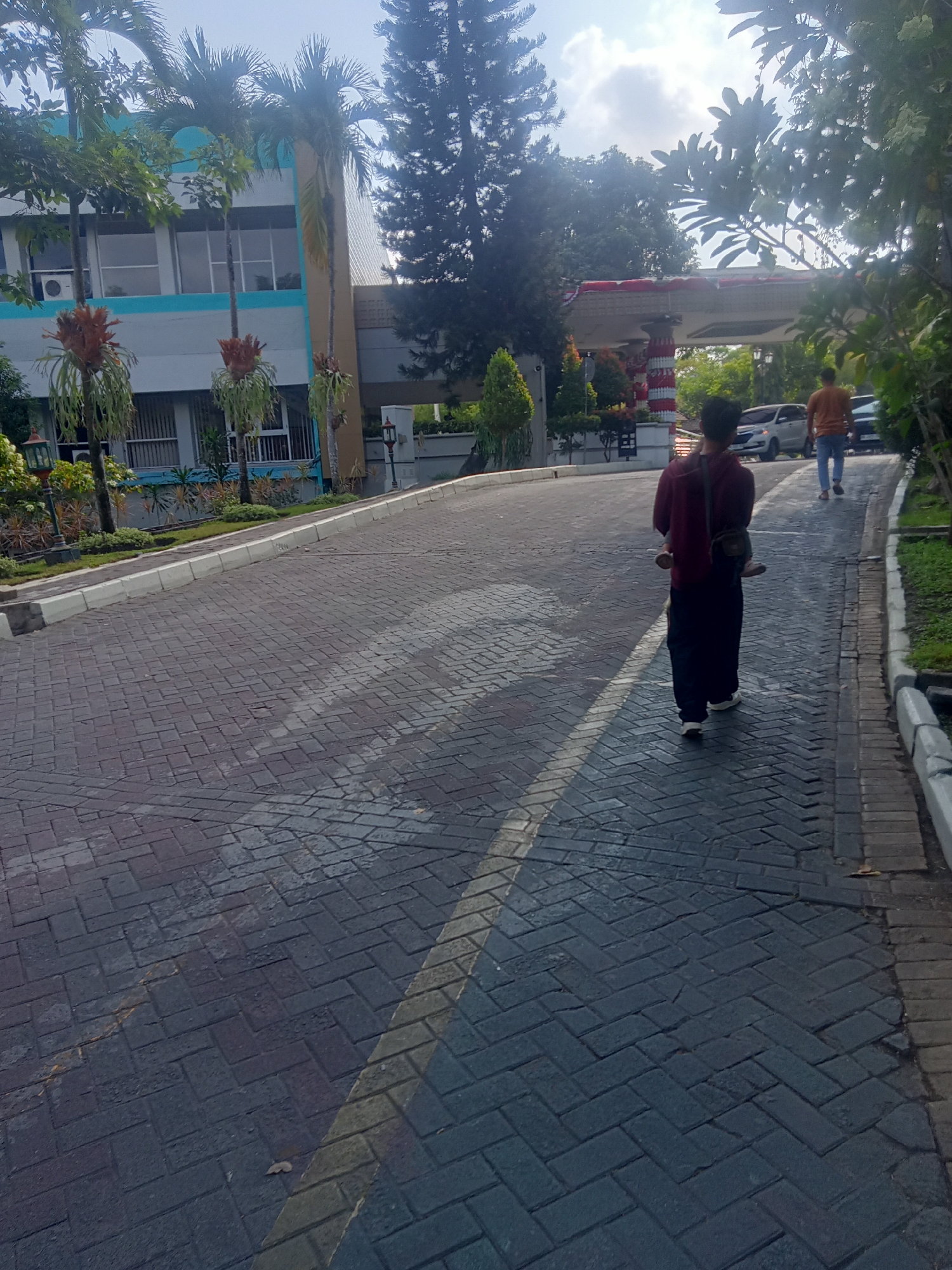 The image shows a street with a person walking away from the camera on the right side of the frame. The person is wearing a dark red hoodie and dark pants, with a bag slung over their shoulder. They are walking on a paved road made of dark grey bricks. There is a yellow line marking the edge of the lane on the right side of the road. Another person in an orange shirt is walking further down the road in the same direction. A silver car is parked on the left side of the road. In the background, there is a modern building with blue and white accents, and palm trees. The scene appears to be in a campus or institutional setting, likely in Depok, Indonesia, given the architectural style and vegetation. The sky is bright and sunny, suggesting it is daytime.