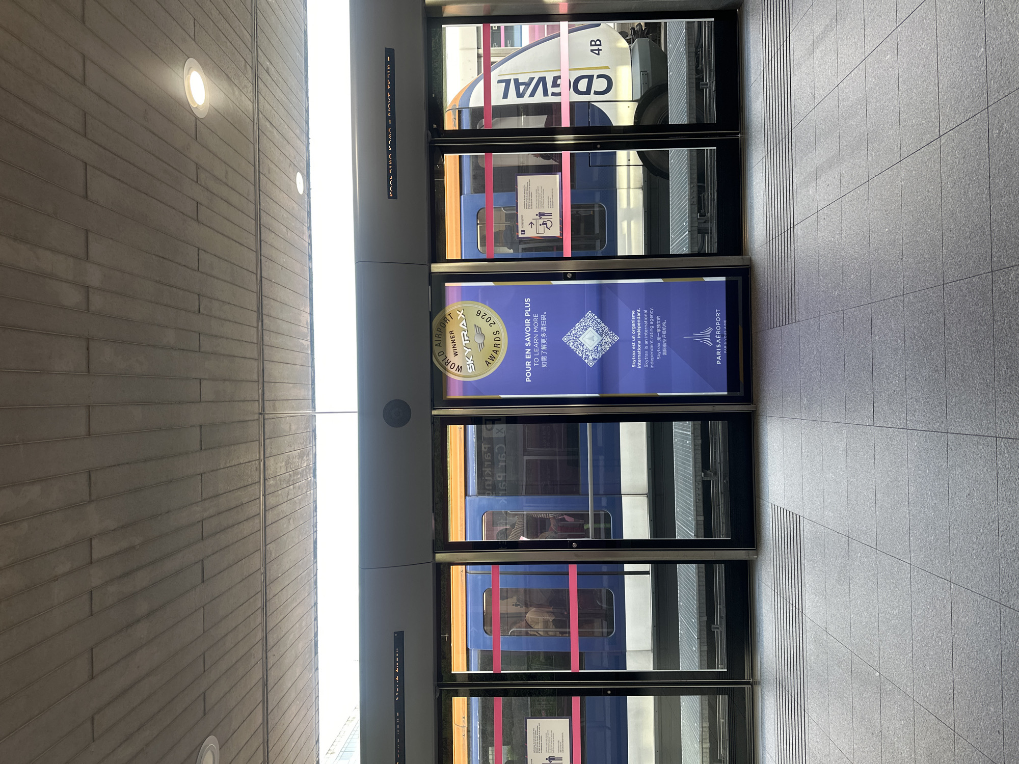 The image depicts a train station platform in Tremblay-en-France, France. A train is visible through glass doors, identified as the CDGVAL, with the car designation "4B" displayed. The platform floor is tiled in gray. Advertisements are affixed to the glass doors, showcasing the "WORLD AIRPORT SKYTRAX AWARDS 2026". Additional text in French and English offers information about Skytrax and Paris Aéroport. The walls are constructed of horizontal stone blocks, illuminated by circular lights.