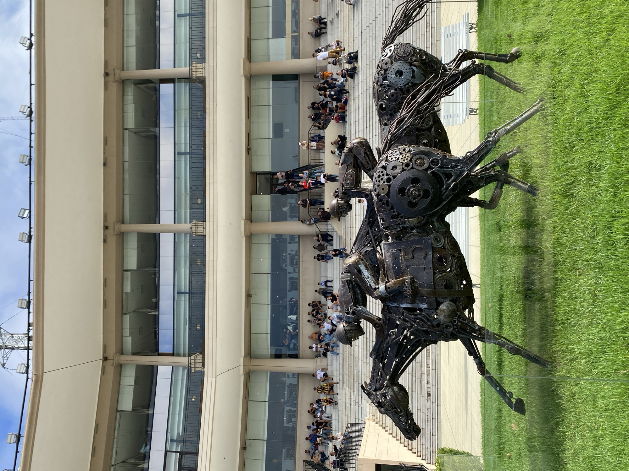 The image shows two metal horse sculptures with riders constructed from various mechanical parts. The sculptures are positioned on a paved surface adjacent to a patch of green grass. Behind the sculptures is a large, multi-story building with visible rows of spectators.
The location is likely the Hipódromo de Palermo (Palermo Racecourse) in Buenos Aires, Argentina, recognizable by the architectural style of the building and the subject matter. Spectators are observing from the building's platforms.