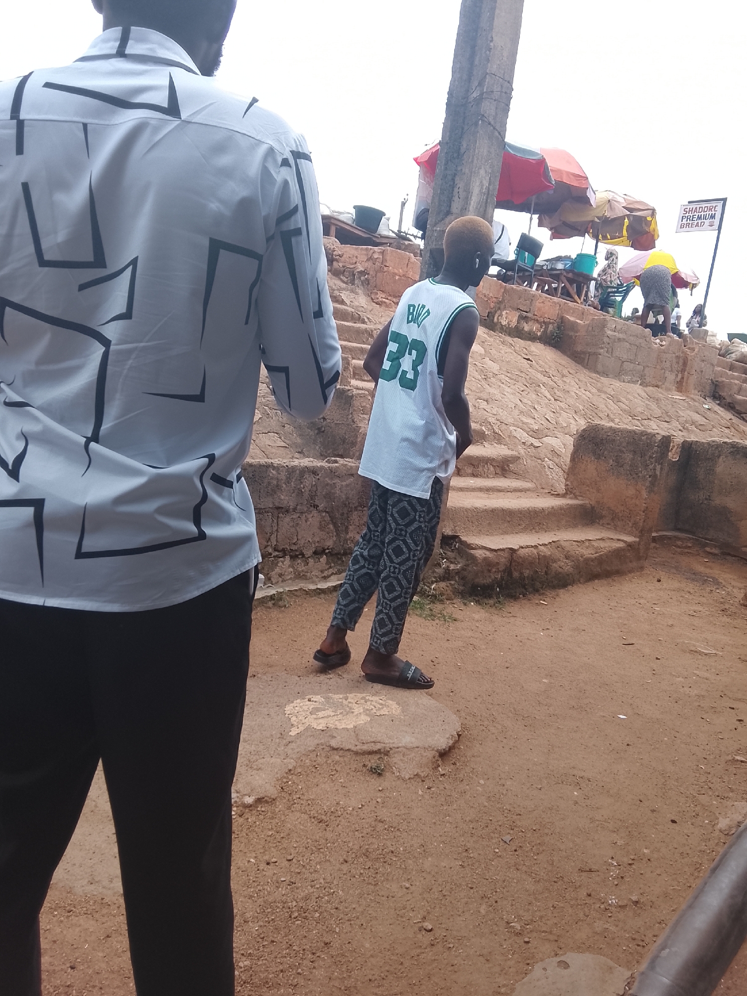 In Jos, Nigeria, an outdoor scene unfolds with a focus on two individuals. The foreground is dominated by the back of a person wearing a white shirt with a geometric black pattern and black pants. To the right, a young person with dyed blonde hair, wearing a white basketball jersey with the number "33" and patterned pants, stands facing away from the camera. This individual appears to be looking towards a set of stone stairs.
In the background, various elements suggest a marketplace or gathering area. Several colorful umbrellas, some red and yellow, others orange and yellow, provide shade. Beneath these umbrellas, people are seated and interacting, with tables and containers visible, indicating commercial activity. A sign is attached to a pole, reading "SHADORE PREMIUM BREAD." The environment is characterized by rough, uneven terrain, with exposed rocks and reddish-brown earth. A concrete pillar stands prominently to the left of the scene. The sky above is overcast.