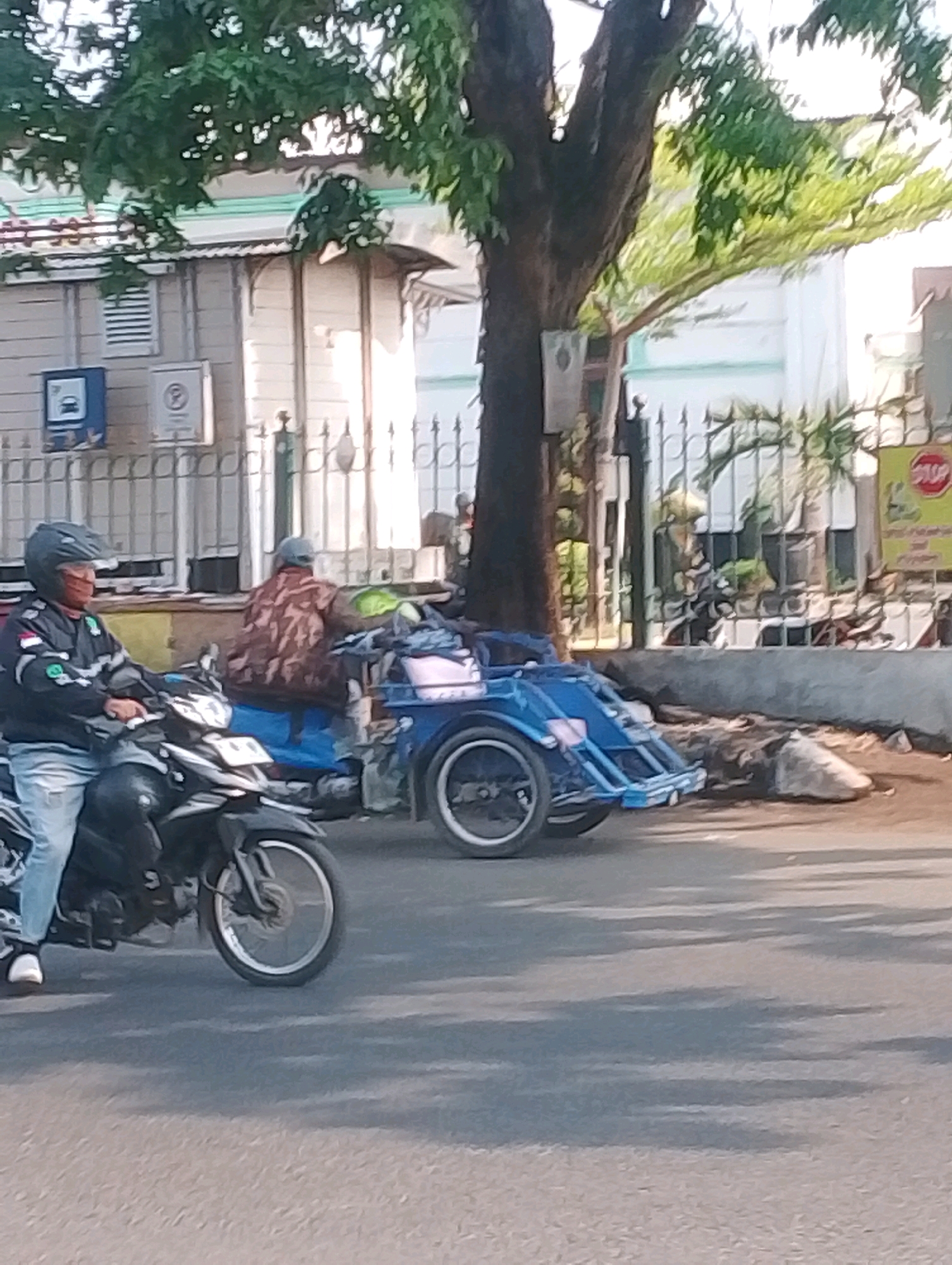 The image depicts a street scene in Probolinggo, Indonesia, under dappled sunlight filtering through trees. In the foreground, a person wearing a black jacket, blue jeans, and a helmet with a face mask is riding a black motorcycle. Behind this motorcycle, a blue motor-rickshaw is visible, with a person wearing a patterned jacket sitting in it. The motor-rickshaw appears to be carrying various items. The background shows a building with light-colored walls and a striped decorative trim, as well as a metal fence. A stop sign is partially visible on the right side of the frame. The road surface is asphalt.