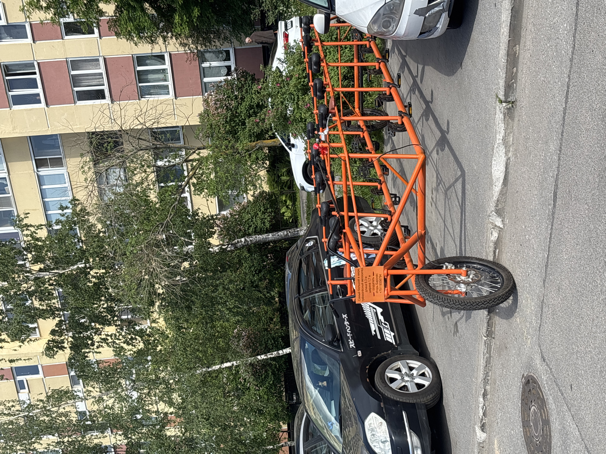A very long, custom-built, orange three-wheeled bicycle (trike) is parked on the side of a paved street in bright daylight. The trike, featuring multiple sets of pedals and seats along its elongated frame, rests against a black car. The black car has "スピードハンターズ" (Speed Hunters) written in Japanese script on its side, along with a logo. Partially visible next to the trike is the front of a white car.
In the background, a multi-story residential apartment building with yellow and reddish-brown sections and numerous windows is visible, partly obscured by green trees. A single person is faintly discernible standing near the building in the distance. The strong sunlight casts clear shadows on the ground and vehicles, suggesting a sunny day, likely mid-morning or afternoon.
A sign attached to the orange trike, written in Cyrillic, provides details: "Самый большой в мире трехколёсный велосипед" (The biggest in the world three-wheeled bicycle), specifying its dimensions as "длина 6,3 м" (length 6.3 m) and "ширина 1,7 м" (width 1.7 m). It also lists the "создатель" (creator) as "Игорь Баронас" (Igor Baronas) and includes a phone number: "+79119356794". The scene captures the trike seemingly on display or temporarily parked.