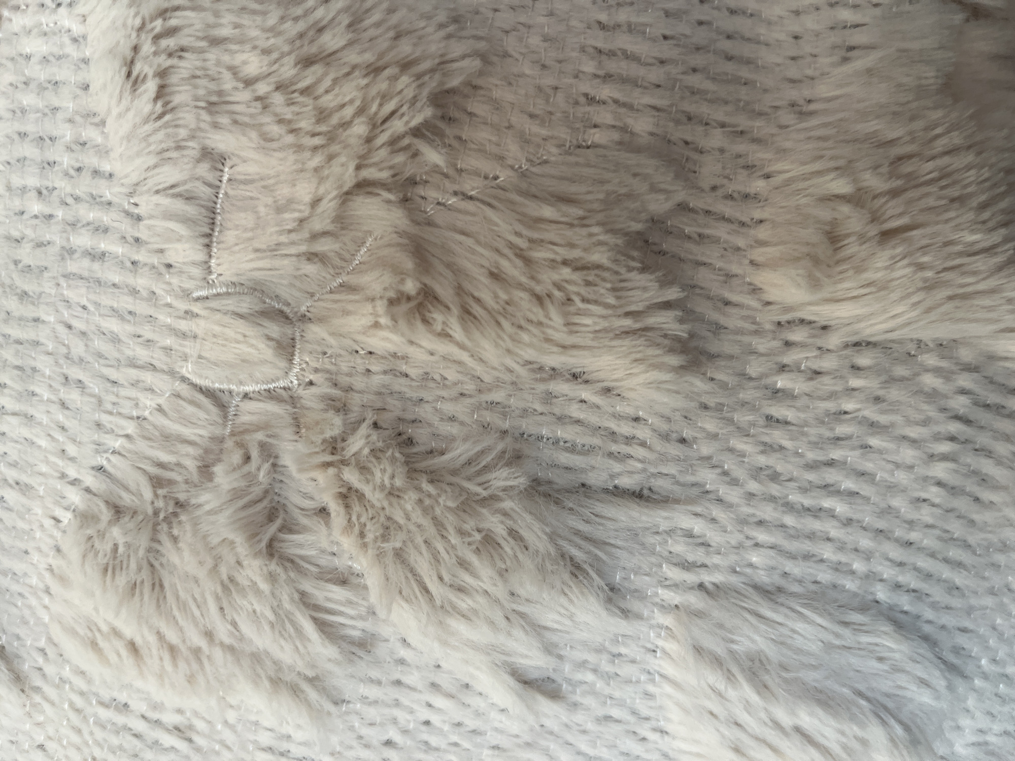 The image shows a close-up view of a textured fabric, possibly a pillow or blanket, with a knitted pattern and fluffy embellishments. The dominant colors are light beige and cream. A decorative stitched pattern, resembling a bow with leaves, is visible in the center of the image, overlaid with the soft, furry material. The knitted background has a subtle sheen, likely due to metallic threads woven into the yarn. There are no people or any discernible elements of the city of Garmisch-Partenkirchen, Germany in the image.