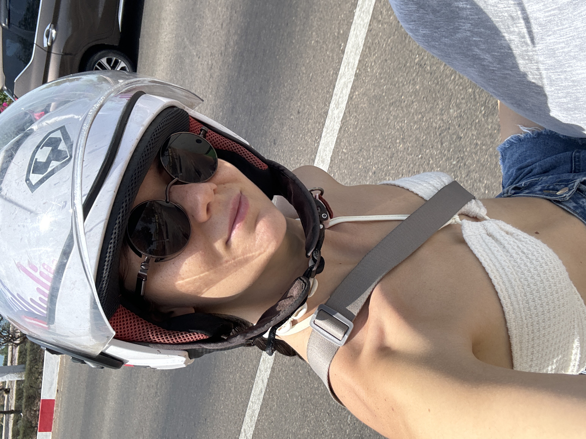 A woman is wearing a helmet and sunglasses while riding a motorcycle in Phu Thuy Ward, Vietnam. She is wearing a white top, jean shorts, and a grey strap. The photo appears to be taken from a high angle. A vehicle is visible in the background.