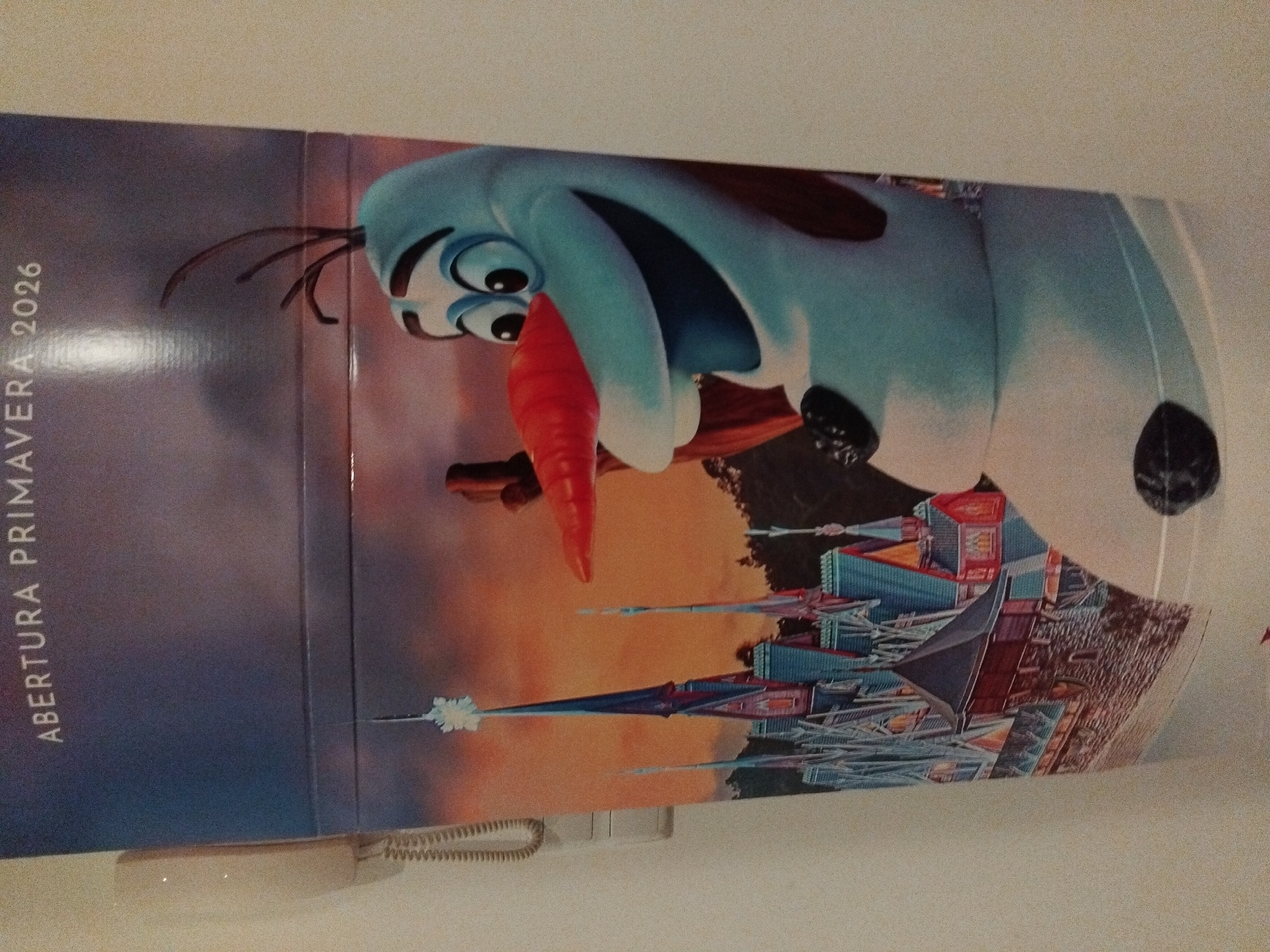 The image shows a large, colorful poster featuring Olaf, the snowman character from Disney's "Frozen," standing in front of a stylized castle. The poster has text at the top that reads "ABERTURA PRIMAVERA 2026." The poster is mounted on a wall. To the left of the poster, a beige wall-mounted telephone with a coiled cord is visible. The overall scene suggests a promotional display or advertisement. The poster itself is the primary subject. The background of the poster depicts a sunset sky and a fantastical castle. Olaf is rendered in his typical blue and white form, with his signature carrot nose and twig-like arms. The poster is predominantly on display in a vertical orientation. The surrounding environment appears to be indoors, with a textured wall. The location in the city of Vila Nova de Famalicão, Portugal, is not directly discernible from the image content.