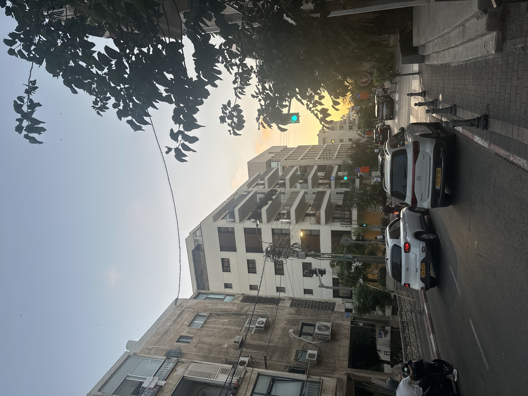 A street scene in Tel Aviv, Israel, captures a moment during sunset. The view is from street level, looking down a moderately wide, paved street lined with multistory buildings. On the left, an older, weathered building with balconies and air conditioning units stands adjacent to a modern, white apartment building with multiple floors and balconies. On the right, a newer, white apartment building dominates the view, its balconies visible against the sky.
The street is occupied by several vehicles. A white SUV is in the foreground on the right lane, its license plate readable. Another white SUV is in the left lane, further ahead. A motorcycle with a rider wearing a helmet is in the left lane, in front of the older building. A few other cars are visible further down the street.
The sky exhibits the warm hues of sunset, with oranges and yellows filtering through the clear sky. Trees with dark green leaves are prominent in the foreground and along the right side of the street, partially obscuring the buildings and sky. Traffic lights are visible, with at least one showing a green light. The sidewalk on the right is paved with cobblestones and bordered by bollards. The overall impression is an urban environment during the late afternoon or early evening, with a blend of older and modern architecture.