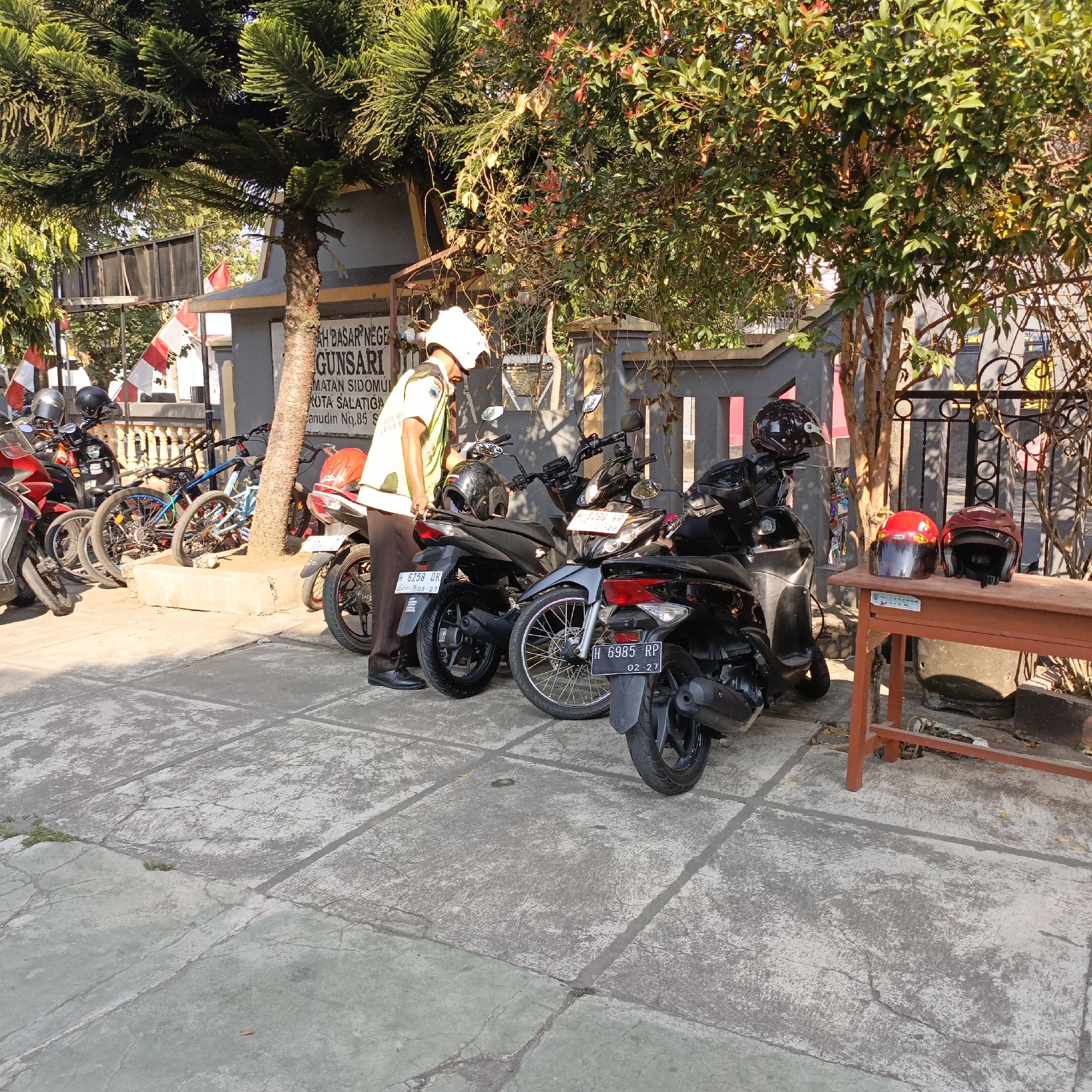 In Salatiga, Indonesia, a person wearing a white helmet and a yellow safety vest stands next to several motorcycles parked on a concrete pavement. The individual appears to be interacting with one of the motorcycles. Several other motorcycles and bicycles are parked in the background, some under the shade of trees. A sign with Indonesian text, possibly indicating a business or institution name such as "TKH. BASAR NEGE GUNSARI", is visible on a building. Red and white Indonesian flags are displayed on poles. The environment suggests an outdoor parking area, possibly adjacent to a building. Two helmets are placed on a wooden table in the foreground.