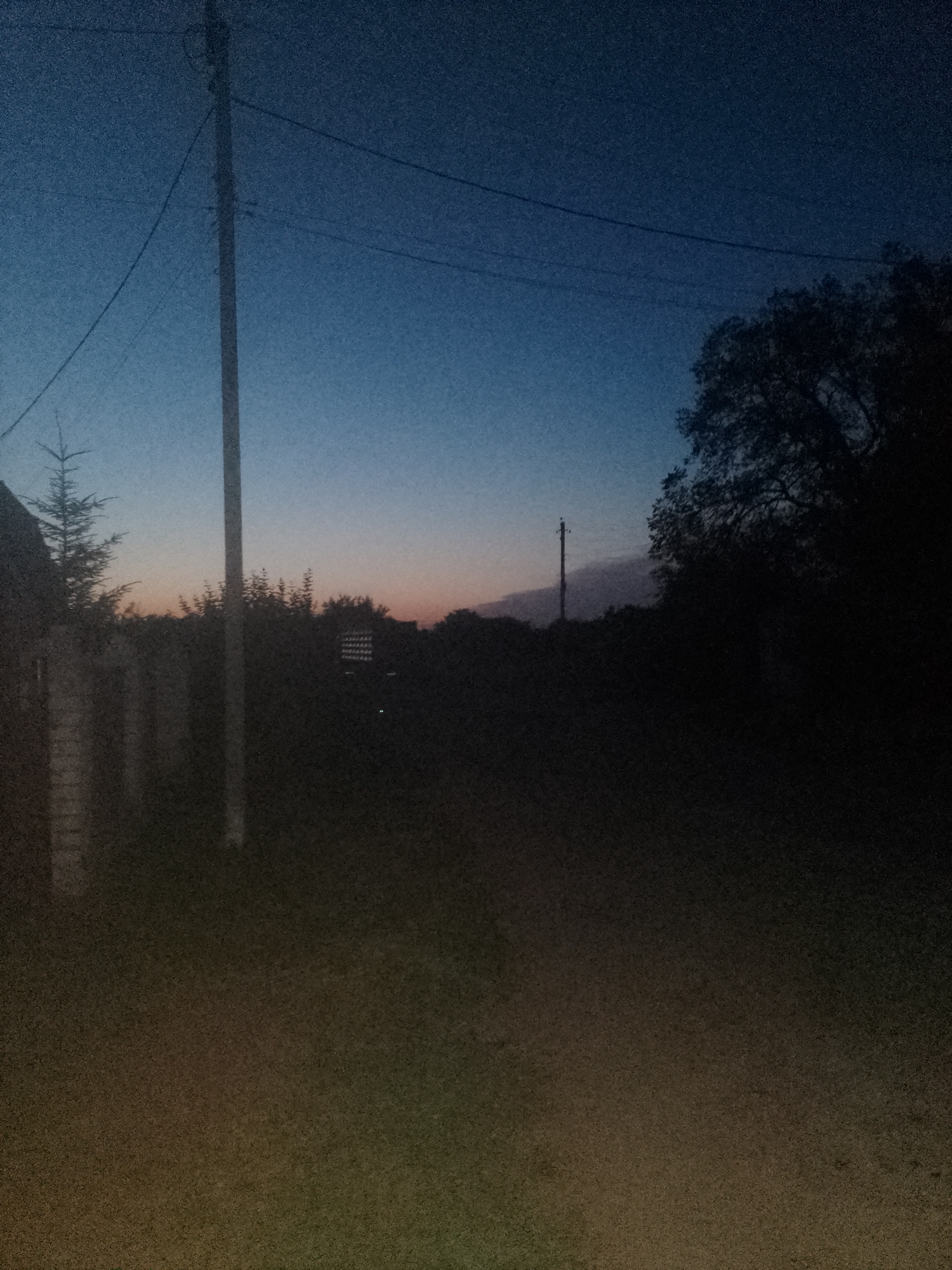 The image depicts a dimly lit outdoor scene at dusk, with a gradient of deep blue to lighter blues and faint orange hues in the sky. A tall, dark utility pole with wires stretches vertically near the center of the frame. To the left, the silhouette of a building or fence is visible. In the foreground, the ground is covered in what appears to be grass or dirt, with a subtle transition of colors from brown to green. To the right, the dark silhouette of a large tree with dense foliage dominates the background. Further into the background, the shapes of other trees and a thin, vertical pole with what looks like a light fixture or signal are discernible. There are no people or discernible animals in the image. The overall impression is a quiet, rural or semi-rural setting at the end of the day. The location is specified as Ustyvytsia, Ukraine.