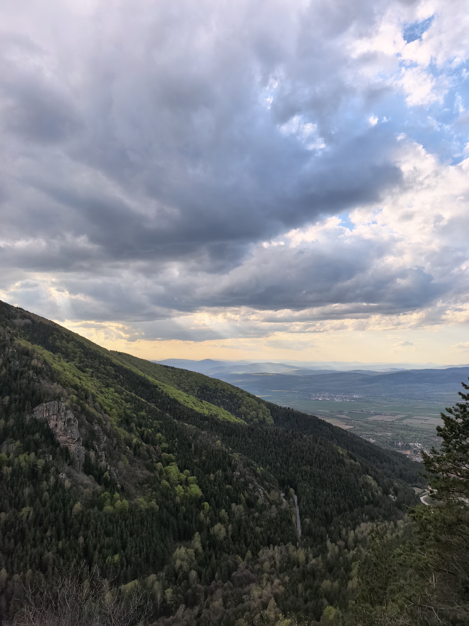 The image displays a vast mountainous landscape, likely near Sapareva Banya, Bulgaria, captured from an elevated viewpoint. A prominent, heavily forested mountain slope dominates the left and center of the frame, showcasing dense coniferous trees mixed with patches of brighter green deciduous foliage, suggesting spring growth. Rocky outcrops are visible on the upper left section of this slope.
Above, the sky is dramatic and partially overcast, filled with large, grey and white clouds. Patches of blue sky are discernible towards the upper right. Striking crepuscular rays pierce through the clouds on the left and center, casting soft light onto the distant landscape and giving parts of the scene a warm, golden hue. This lighting suggests either a late afternoon or early evening setting.
Below the immediate forested slope, a wide valley unfolds, containing agricultural fields and a small town or village with buildings visible. Further in the distance, successive layers of hills and mountains fade into a hazy, atmospheric perspective. A winding road can be faintly seen descending the mountain on the lower right, leading towards the valley.
No people or specific activities are visible within the frame, and there is no discernible text. The scene evokes a sense of natural majesty and the serene beauty of a mountainous region under a dynamic sky.