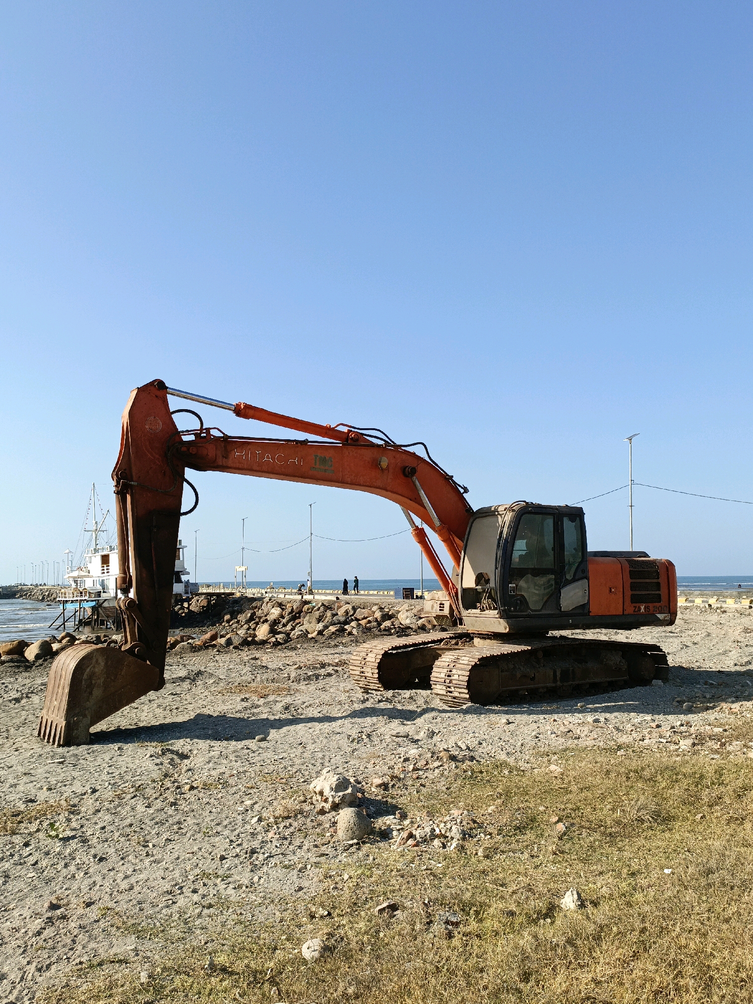 An orange Hitachi excavator, model ZX 200, sits idle on a sandy, rocky construction site next to a body of water, likely a harbor or coastline, in Bulukumba, Indonesia. The excavator's large bucket is lowered to the ground, and its boom extends over the terrain. In the background, a white boat is docked, and a pier with lampposts extends into the water. Two figures are visible walking on the pier. The sky is clear and blue, and the sun casts shadows on the ground. The scene appears to be a construction project or port development.