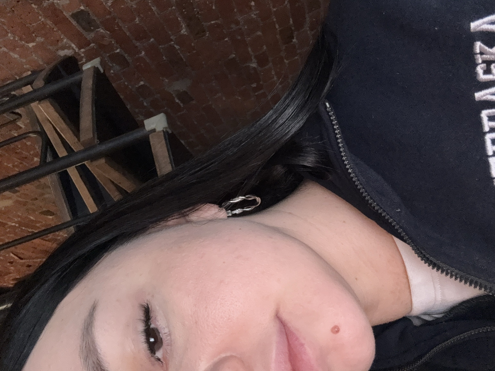 A close-up, angled selfie shows a woman's face, partially obscured by black hair. She has fair skin, dark eyes, and a mole near her lip. She wears a twisted silver earring. She is wearing a black zip-up jacket with white lettering. A portion of a brick wall and metal staircase are visible in the background. The location in Moscow, Russia cannot be confirmed from the provided image.