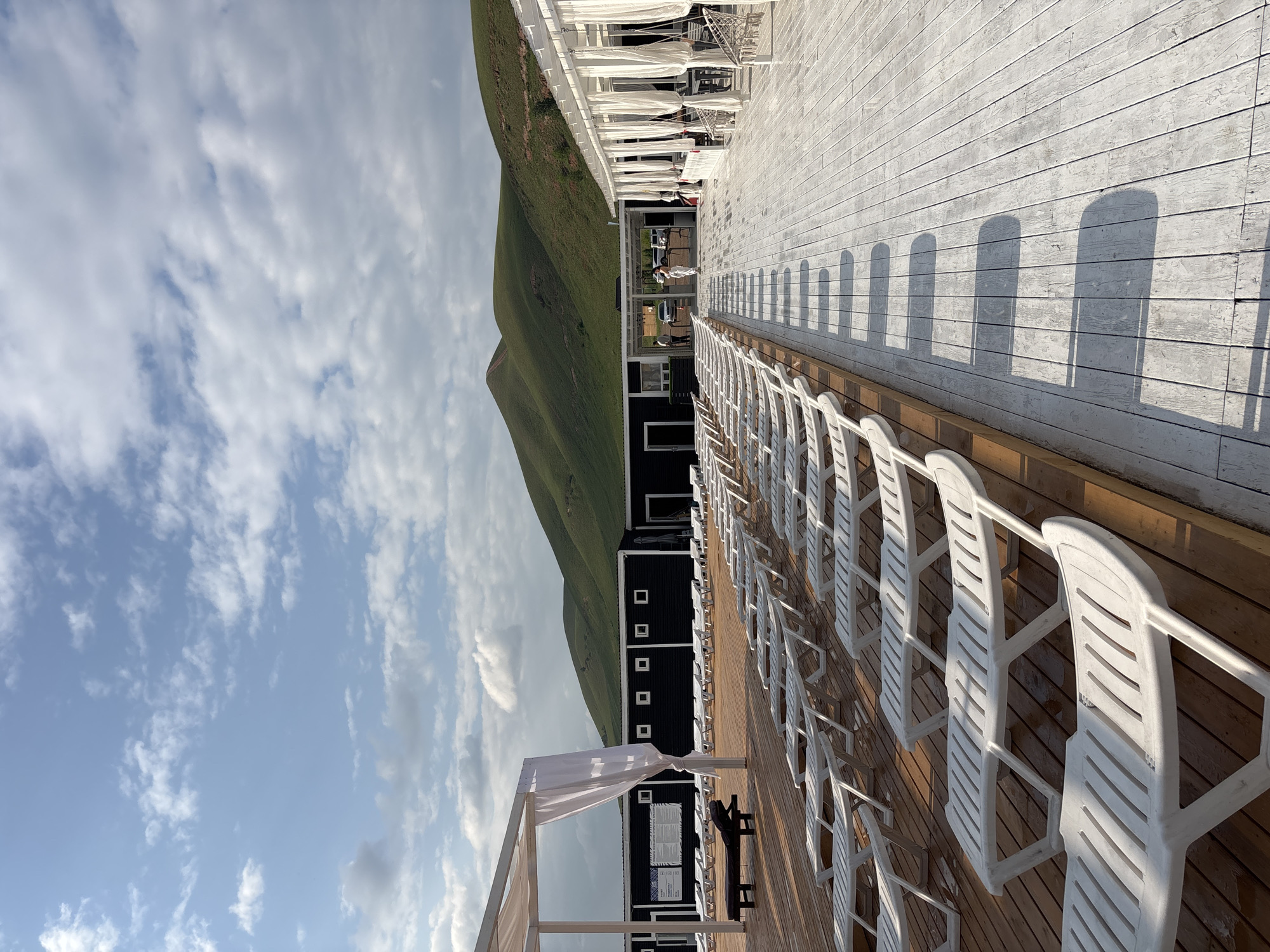 The image depicts an outdoor resort area, likely a beach or poolside location, with rows of white lounge chairs on wooden decks. The resort features white structures with draped fabric, providing shade and a resort-like ambiance. In the background, green, rolling hills dominate the landscape under a partly cloudy sky.
The scene appears to be a resort in the vicinity of Дрокино, Russia, given the presence of natural, hilly terrain characteristic of the region. The resort itself consists of several structures: one is a long, dark building with numerous windows, likely housing amenities or rooms. Another is a white, open-air structure with white curtains, possibly a dining or lounge area. The wooden decks are clean and well-maintained, with lounge chairs neatly arranged. Some people are visible in the background, appearing to be staff or guests, interacting near the entrance of the dark building. The overall impression is a tranquil, scenic resort setting, ready for visitors.