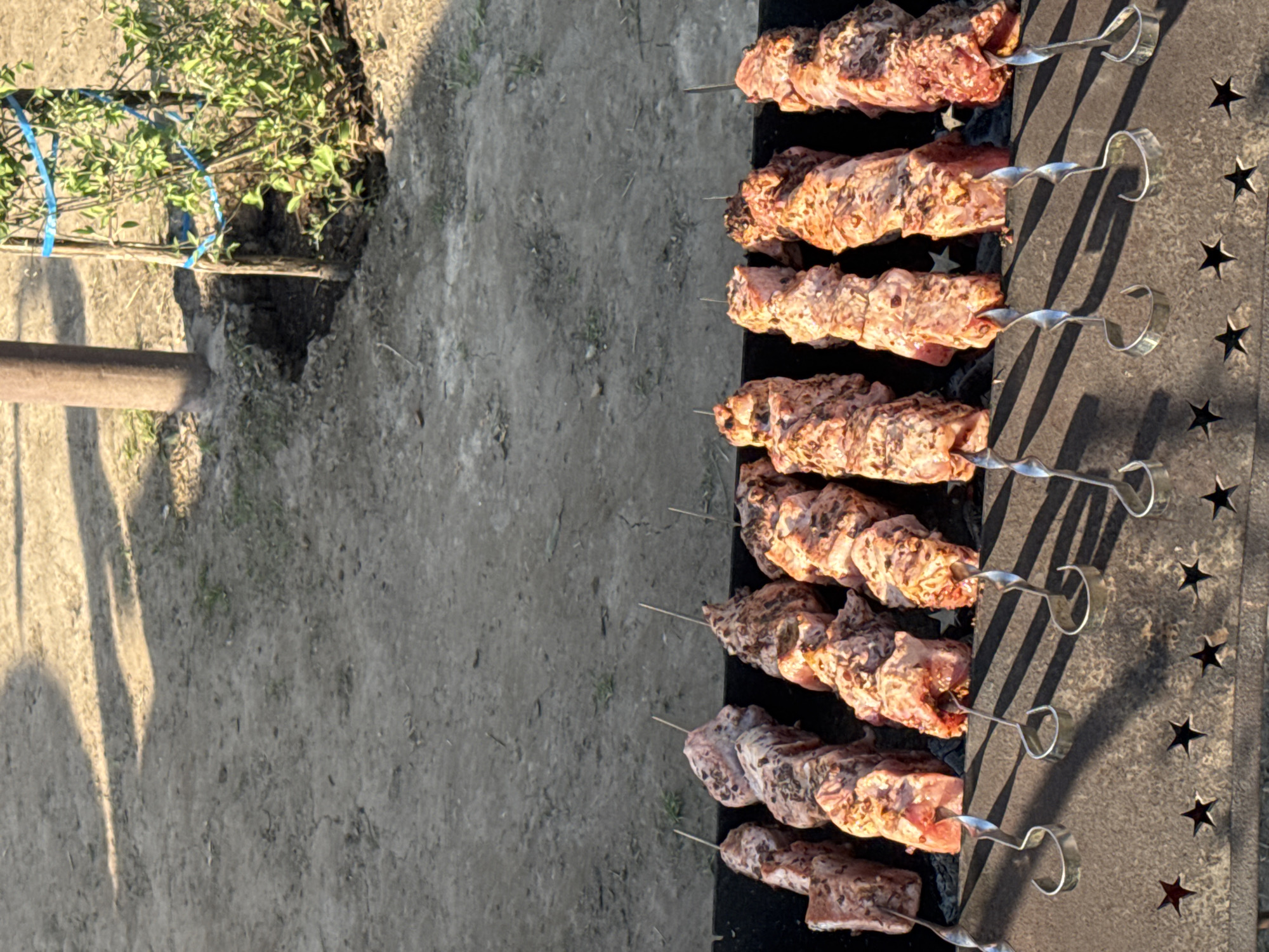 The image shows meat skewers cooking on a grill with star-shaped cutouts, sitting on a dirt surface. In the background, a small tree is visible. The location is likely an outdoor area near Krasnokamsk, Russia.