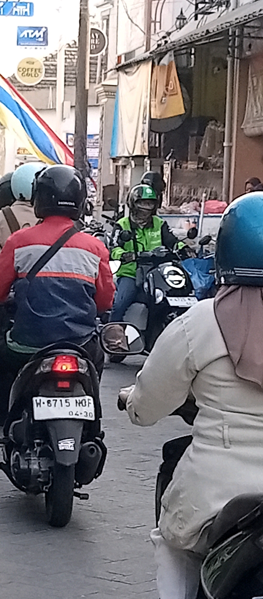 The image captures a street scene with several motorcycles and riders in Wadungasri, Indonesia. In the foreground, a motorcycle with a black body and a red taillight is visible from the rear. Its license plate reads "H - 6715 NQF" with "04-30" below it. The rider of this motorcycle is wearing a red, white, and blue jacket and a black helmet. To the right of this motorcycle, another rider is seen from the side, wearing a light-colored jacket and a hijab, and is on a motorcycle with a blue helmet. Further in the background, a motorcycle rider in a bright green jacket and helmet, identifiable as a Gojek driver, is also on a motorcycle. Behind them, more motorcycles and riders are present, along with buildings lining the street. The environment appears to be an urban setting with shops and signage, including an ATM sign and a "COFFEE GOLD" sign, suggesting commercial activity. The road is paved with what looks like cobblestones or interlocking pavers.