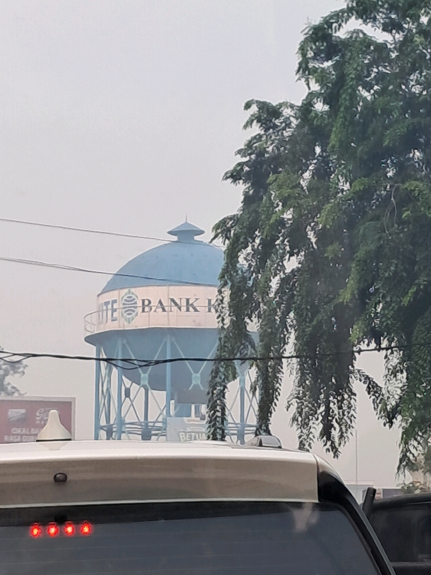 The image depicts a water tower with the logo "BANK K" and a circular emblem visible. The tower is blue with a lighter colored band around its base. A tree with dense green foliage partially obscures the view of the tower. In the foreground, the roof of a vehicle is visible, with four red lights illuminated at the rear. The sky is overcast and hazy. The scene appears to be in an urban or semi-urban environment in Pontianak, Indonesia, suggested by the presence of the bank logo which is likely related to Bank Kalbar, a regional development bank in West Kalimantan, Indonesia.