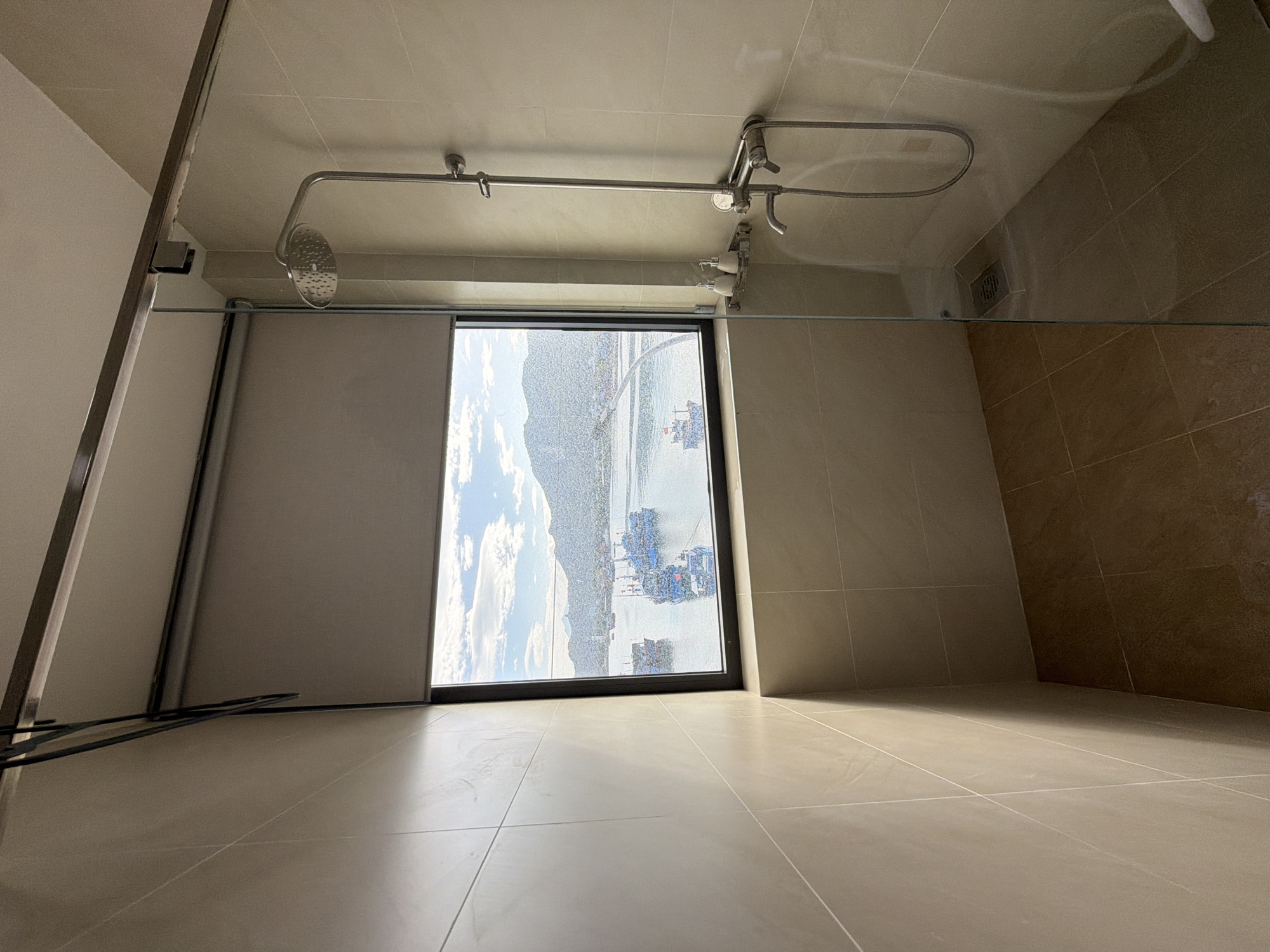 The image captures an interior view of what appears to be a modern bathroom or shower stall, taken from a low angle looking upwards. The floor and walls are covered in light beige tiles with visible grout lines, while a section of the wall to the right features darker brown tiles.
In the upper central part of the frame, an overhead shower fixture is visible, consisting of a metal pipe leading to a circular shower head, along with a separate handheld shower on a rail and control knobs. A transparent glass shower screen extends from the ceiling downwards, partially reflecting the ceiling and an apparent floor drain. On the left side of the shower area, a rolled-up gray blind partially covers the window. Below the blind, near the floor, a dark, linear metal object, possibly a towel rail or drying rack, is positioned.
The dominant feature of the image is the large window in the center, offering an expansive view of an outdoor scene. Through the window, a body of water, likely a sea or bay, is visible, populated by numerous boats, many of which appear to be fishing vessels with distinctive blue elements. Beyond the water, a mountainous or hilly landscape stretches into the distance under a bright, partly cloudy sky. White, fluffy clouds are scattered across the light blue sky.
The scene depicts daylight conditions, suggesting fair weather. There are no people visible, and no specific activity is taking place. There is no visible text in the image. The coastal view with numerous fishing boats is consistent with the provided location context of Ninh Chu Ward, Vietnam.
