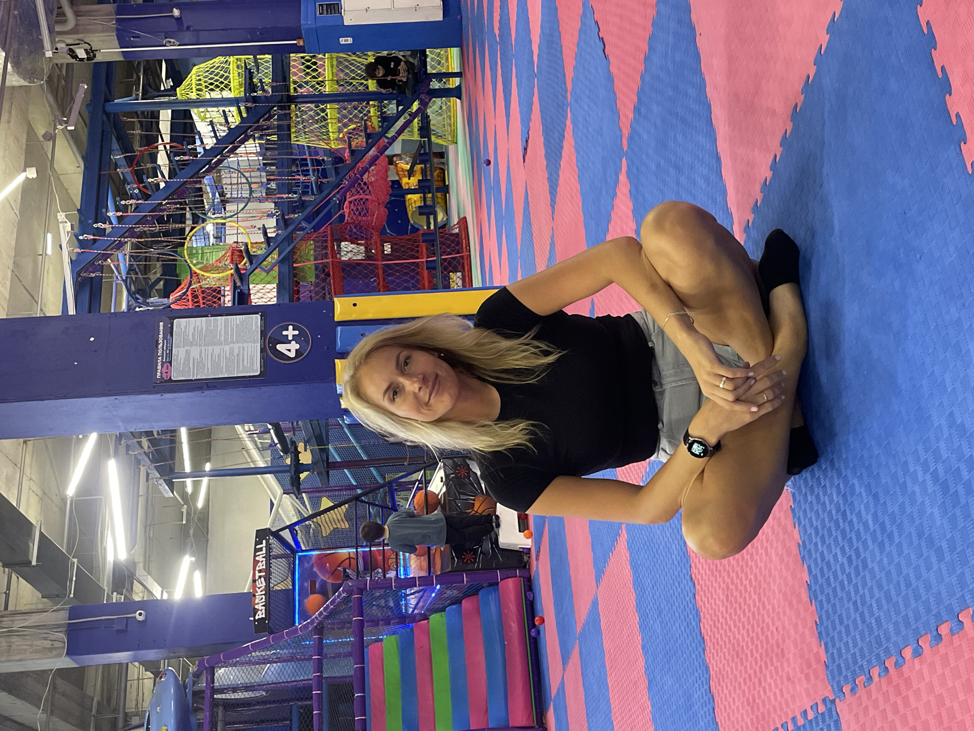 A woman sits cross-legged on a blue and pink puzzle-piece mat in an indoor playground. She wears a black t-shirt, gray shorts, and black socks. The playground features colorful climbing structures, a basketball area with balls, and various play zones constructed with metal framing, netting, and soft padding. The location is likely Sochi, Russia, considering other metadata.