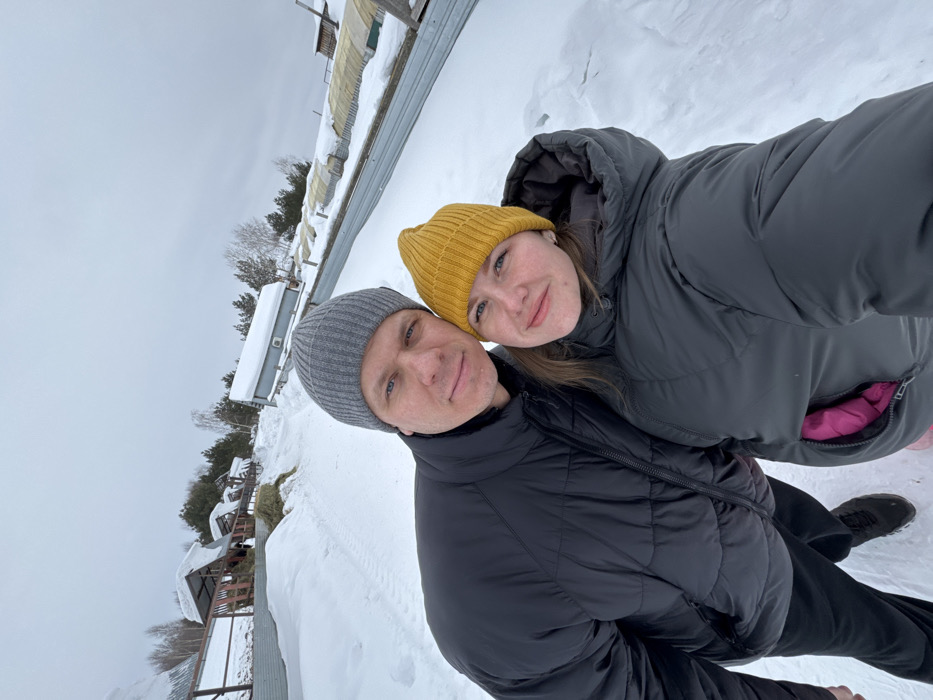 This image depicts an adult male and an adult female posing for a selfie in a snow-covered outdoor environment in Crokhovo, Russia. The male, positioned on the left, has light skin, blue eyes, and wears a grey knitted beanie and a black puffer jacket. His left arm is around the female. The female, positioned on the right, has light skin, light-colored hair partially visible, and wears a bright yellow knitted beanie and a dark grey puffer jacket; a pink garment is visible within an open section of her jacket. Both individuals are looking directly at the camera with neutral to slight smiles.
The background is entirely covered in snow under an overcast sky. To the left, an upward sloping area features a corrugated metal fence, beyond which are several wooden structures with sloped, snow-covered roofs, resembling animal shelters or feeding troughs containing hay. Coniferous and deciduous trees are visible further back. To the right, another corrugated metal fence parallels the foreground, behind which are several elongated, low-profile structures covered in snow, consistent with greenhouses or poly-tunnels. A small, light-colored building with a dark roof and an external air conditioning unit is also present on the right. The scene indicates a winter day in a rural or agricultural setting.