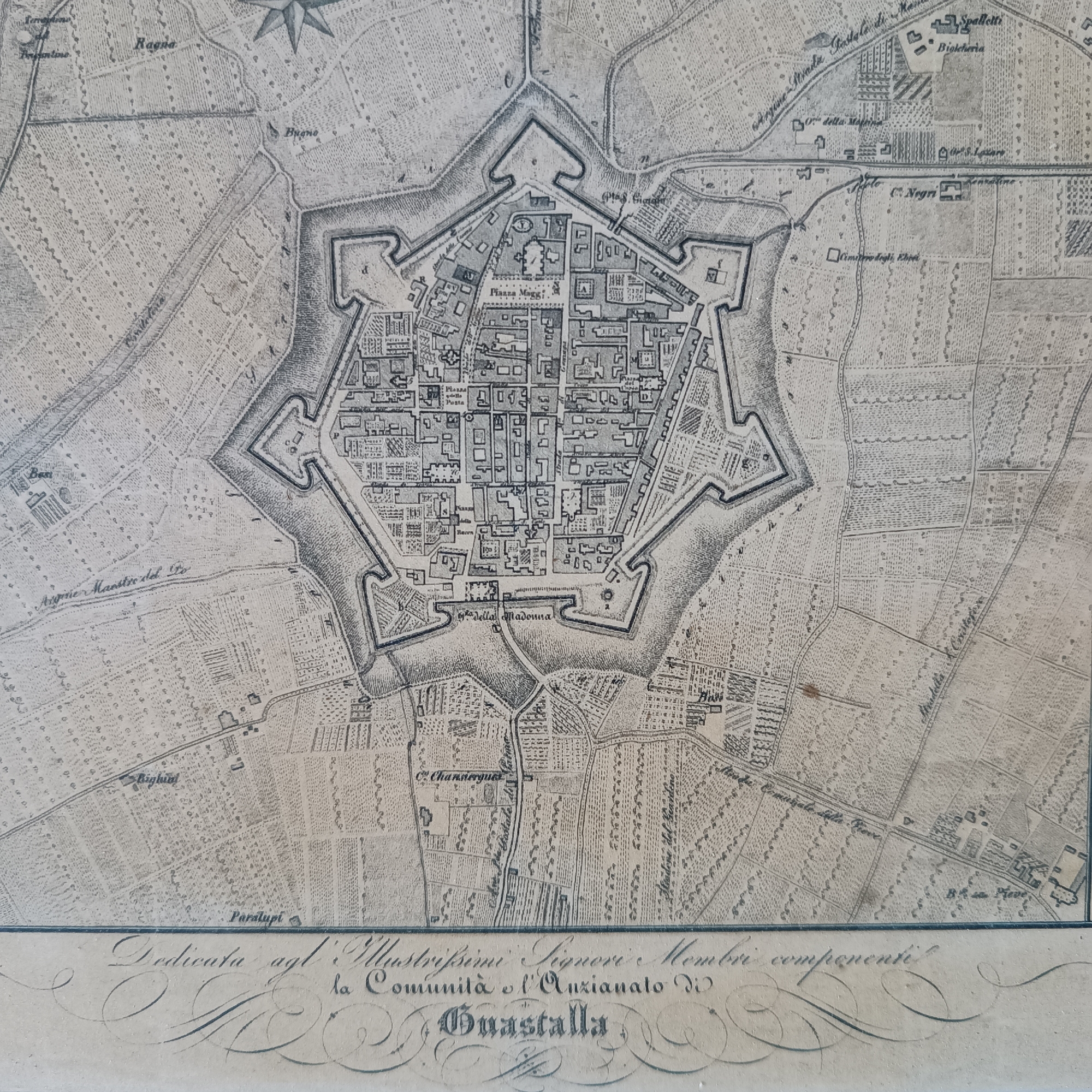 The image is an old map depicting the city of Guastalla, Italy. The city is shown as a star-shaped fortification surrounded by fields, canals, and roads. Within the city walls, a structured layout of streets and buildings is visible, including a "Piazza Maggi" (main square) and "Piazza della Posta". Outside the city walls, various buildings and landmarks are labeled, such as "C. Negri", "Spalletti", "Biolcheria", and "Cimitero degli Ehrei" (Cemetery of the Jews). The text at the bottom indicates the map is dedicated to the "Illustrissimi Signori Membri" (Illustrious Members) of the "Comunità e l'Anzianato di Guastalla" (Community and the Elders of Guastalla). Mezzana Sant'Antonio is not specifically indicated on the map.