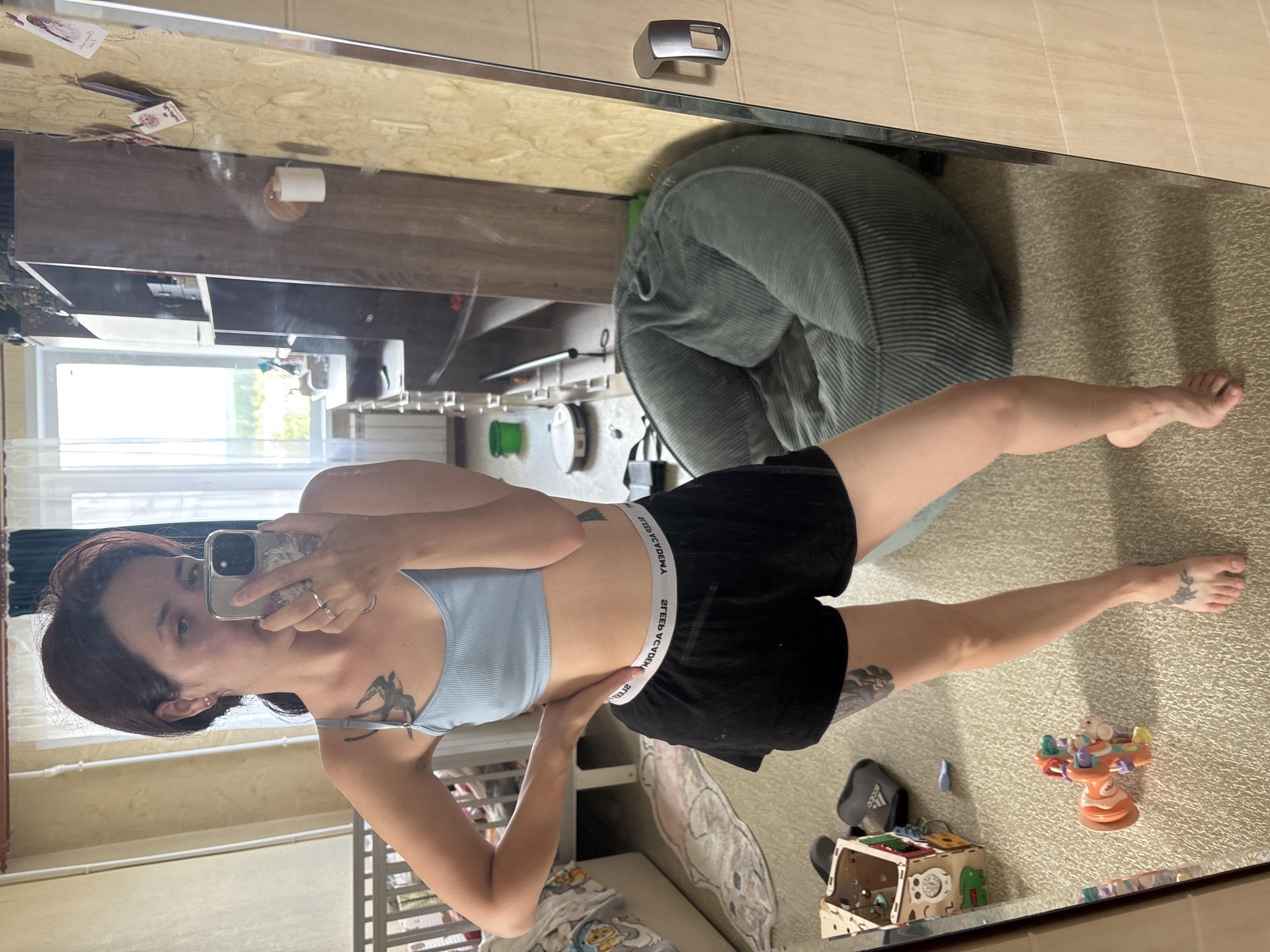 A young woman stands in a bedroom, taking a mirror selfie. She is wearing a light blue bralette and black shorts. A tattoo of a bird is visible on her left shoulder, and another tattoo is on her right thigh. Her left hand rests on her hip, and she holds her phone in her right hand.
The room appears cluttered, with a crib in the background, a child's toy on the floor, and various items on shelves. A large, green beanbag chair is visible in the reflection of the mirror, and a robot vacuum cleaner is on the floor. The room has light-colored walls and a pale carpet. The window in the background shows a view of greenery and trees, suggesting a suburban or semi-rural setting. The presence of a crib and children's toys indicates that the room likely belongs to a family with young children. There is no direct indication of the city of Naberezhnye Chelny, Russia, within the image itself.