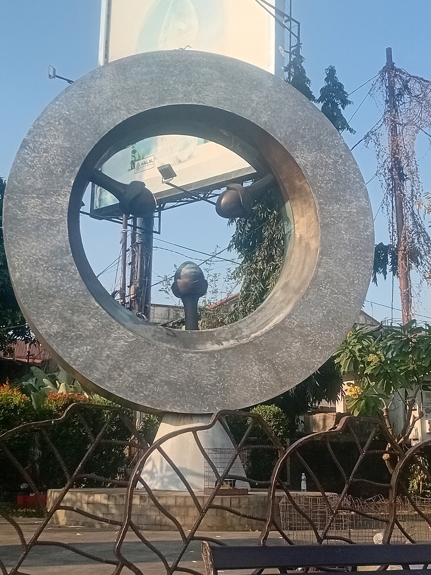 The image depicts a large, circular concrete sculpture in an outdoor setting. The sculpture has a rough, textured surface. Through the center of the sculpture, several objects are visible: a billboard with a "HALAL INDONESIA" logo, trees, electrical poles with wires, and some greenery. In the foreground, there is a decorative metal fence and some plants. The sky is clear and blue, suggesting it is daytime. The scene appears to be in a public space, possibly a park or along a road, given the presence of the billboard and the urban elements. The specific location in Kudus, Indonesia, is indicated by the "HALAL INDONESIA" logo on the billboard, which is a certification relevant in Indonesia.