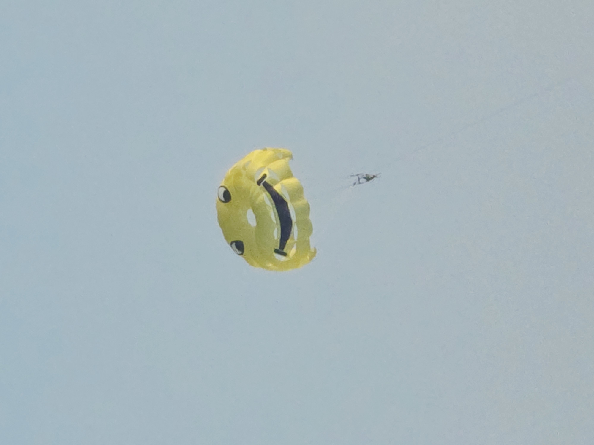 A yellow parachute in the shape of a smiley face is descending against a pale blue sky. A person is suspended beneath the parachute by thin lines. The parachute is the primary subject, occupying the upper half of the frame. Its features, including two circular eyes and a curved mouth, are clearly visible. The person, a dark silhouette, is small in comparison to the parachute. The background is uniformly the sky, suggesting an outdoor activity. The image appears to have been taken during daylight hours.
