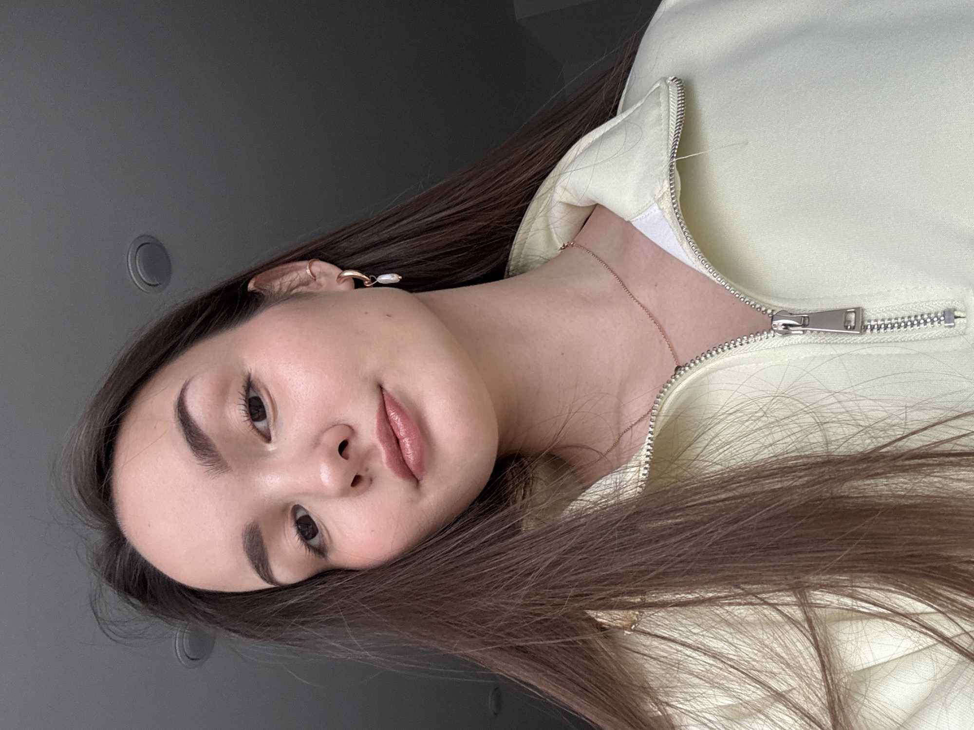 The image shows a young woman with long, dark brown hair posing in what appears to be an indoor setting. She is wearing a light yellow zip-up jacket over a white top and a thin gold chain necklace. She has brown eyes, well-defined eyebrows, and appears to be wearing subtle makeup. Her hair is spread out around her face and shoulders. The background is a plain gray wall with a circular ceiling fixture visible in the upper left corner. There is no information in the image to confirm its location within Chaikovsky, Russia, or any specific location.