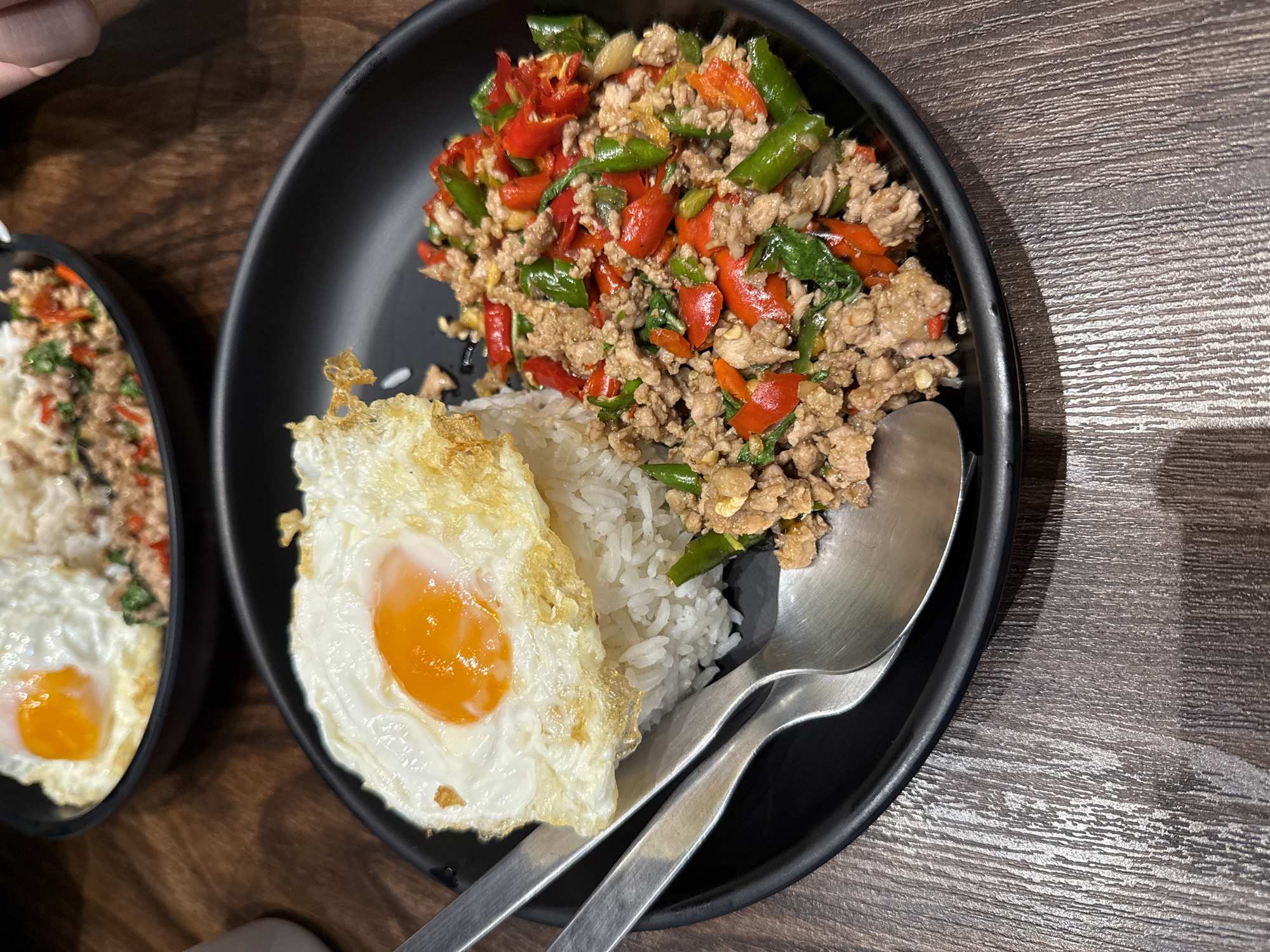A close-up, high-angle shot captures a plate of Thai basil stir-fry with minced meat and a fried egg, served over rice. The dish is presented on a black plate with two silver spoons resting on the side. The background reveals a wooden table and another plate of food, suggesting a dining setting in Bangkok, Thailand. The food appears to be a popular street food dish, commonly found in local eateries and markets across the city. The lighting is natural and soft, highlighting the textures and colors of the food. The overall impression is one of a delicious and satisfying meal, ready to be enjoyed.