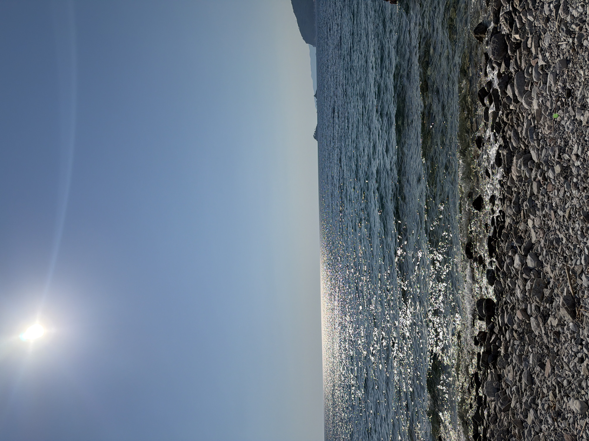 The image depicts a sunlit Adriatic Sea coastline in Golubovići, Montenegro. The foreground is a pebble beach with smooth, grey and white stones of various sizes. The water is a deep blue, with the sun's bright reflection creating a shimmering pathway across its surface. Gentle waves lap at the shore. In the distance, hazy mountains form the horizon, with a small island visible to the left. The sky is clear and bright blue, with the sun positioned in the upper left quadrant, casting a strong glare. A portion of a person's head and shoulder are visible in the lower right corner of the frame, suggesting they are in the water.