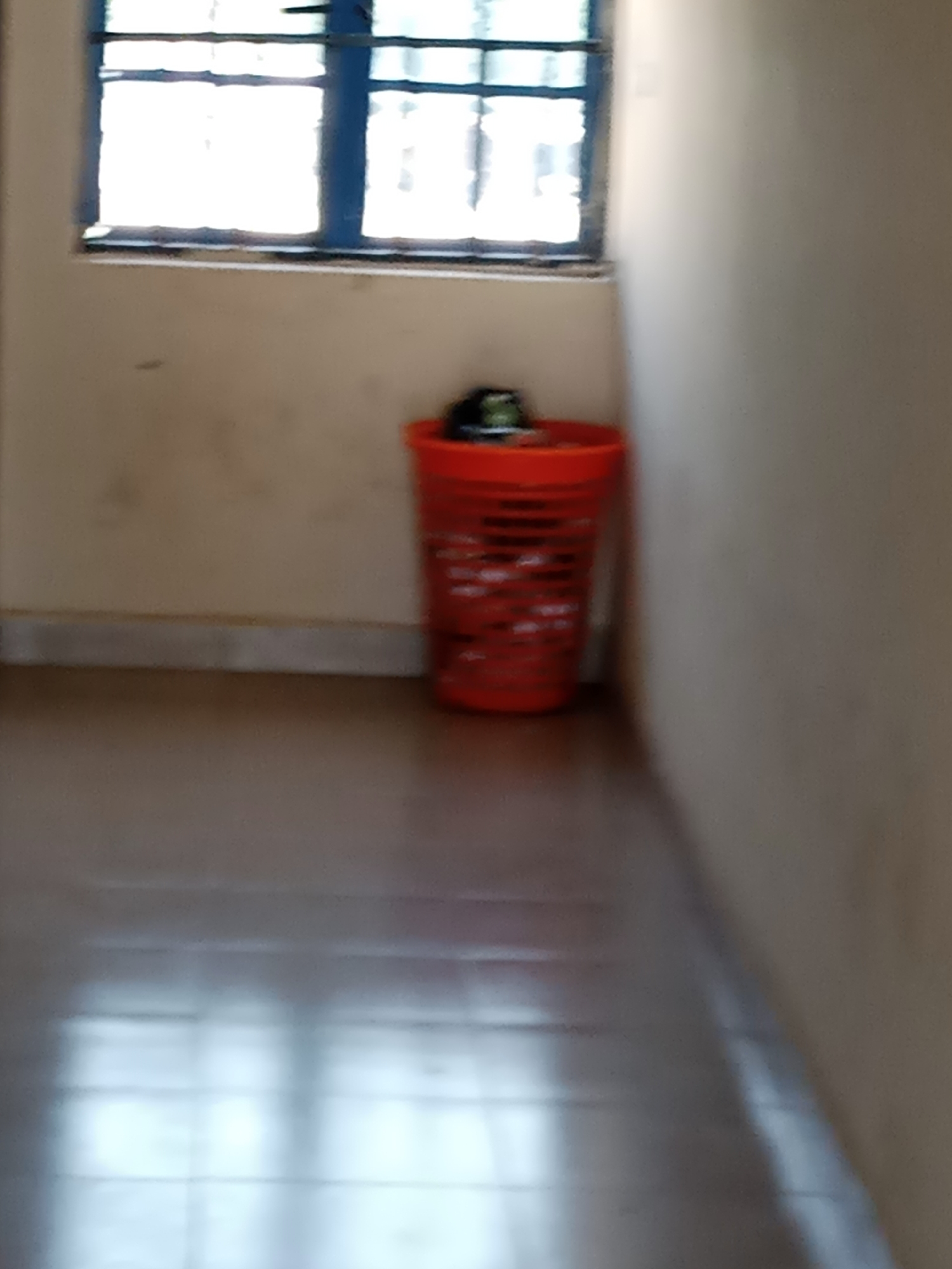 In Jalingo, Nigeria, an indoor scene features a window with blue framing and white panes, possibly indicating a residential or institutional setting. Adjacent to the window, a bright orange plastic basket sits on the floor, containing a dark object, possibly a helmet. The walls are a neutral, light beige color, and the floor appears to be tiled with a light brown hue, reflecting light from the window. The overall impression is one of a simple, functional interior space.
