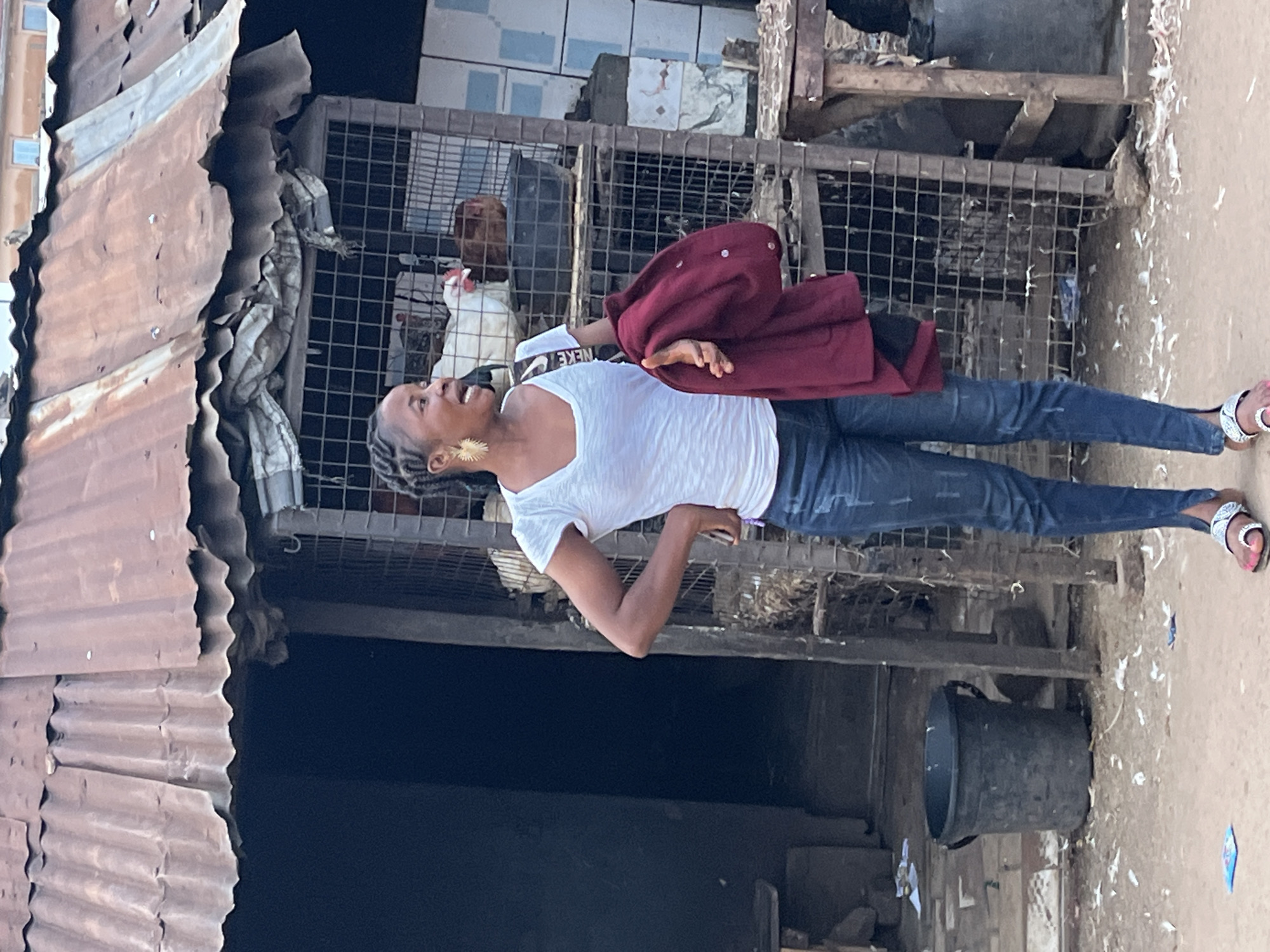 A full shot captures a Black woman standing in what appears to be a market stall in Jos, Nigeria. The woman is wearing a white V-neck t-shirt, ripped blue jeans, and white sandals with a patterned strap. She has her hair braided and pulled back, and she is wearing large, gold, starburst-shaped earrings. She is holding a burgundy jacket or sweater in her right arm and has her left hand on her hip. Her mouth is open as if she is speaking or singing. Behind her, a metal cage contains live chickens. The stall has a corrugated metal roof and a wooden structure. The ground is dirt and scattered with debris, including white feathers and some paper. To the left of the woman, a large black plastic bucket sits on the ground. The background shows a dark entrance to a building and some tiled walls.