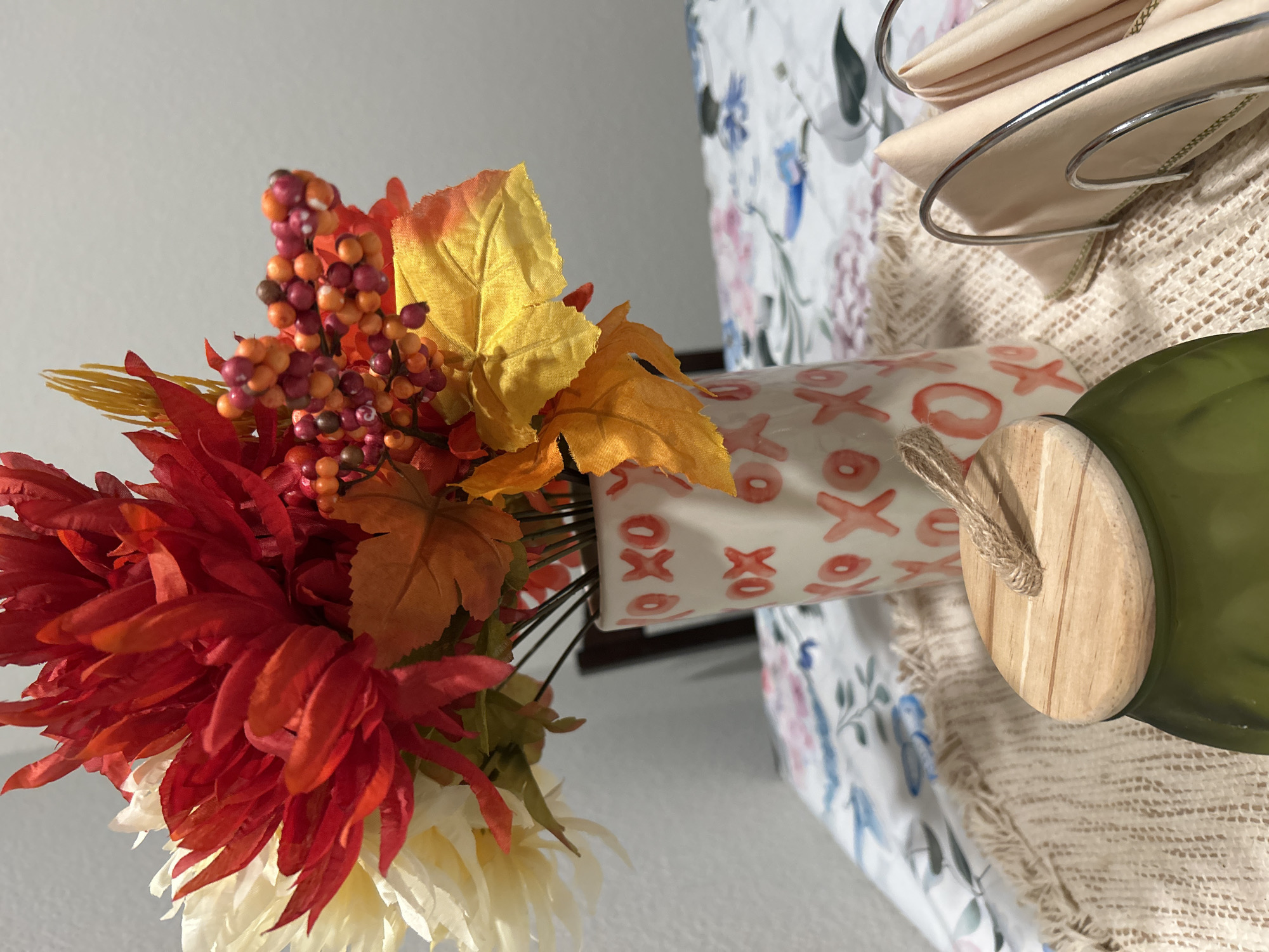 An indoor, eye-level medium shot captures a fall-themed decorative arrangement on a table. The primary subject is a bouquet of artificial flowers and leaves in a white ceramic vase adorned with red "X" and "O" patterns. The flowers are predominantly red and cream, with accents of autumnal berries and faux leaves in shades of orange and yellow. The vase sits on a woven placemat, which in turn is placed on a tablecloth with a floral print featuring pink and blue flowers on a white background. To the right of the vase, a silver napkin holder contains folded light peach napkins. In the foreground, the lid of a green glass jar with a wooden top and a jute rope handle is visible, resting on the placemat. The background consists of a plain, off-white wall, suggesting an indoor setting, possibly a dining room or living area. The lighting is soft, creating a warm and inviting atmosphere. The scene is devoid of people or any discernible action. The image does not provide any information about its location in Colorado Springs, USA.