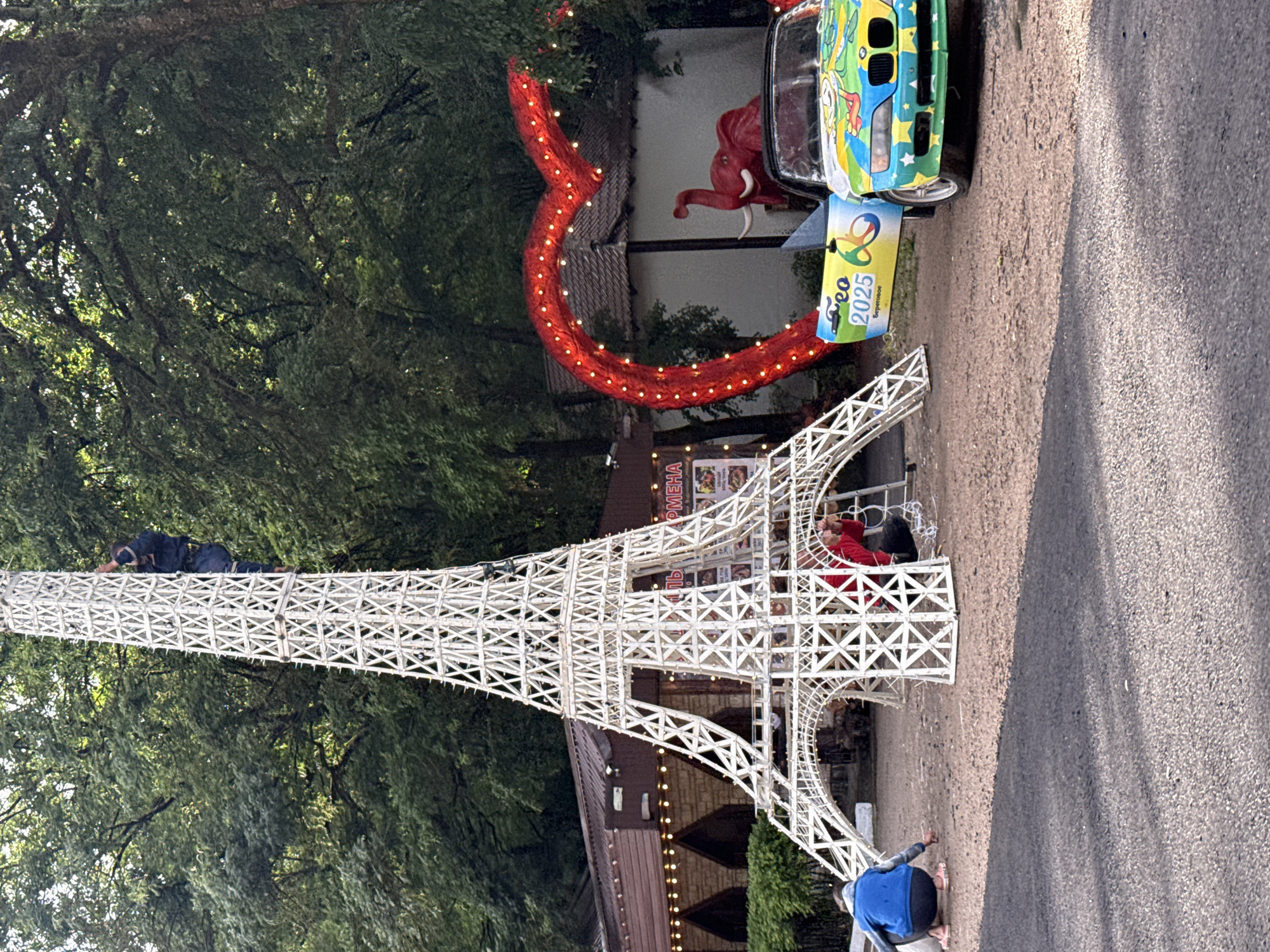 In an outdoor, possibly park-like setting with trees in the background, a white, Eiffel Tower-shaped structure is being assembled or decorated. One person is visible climbing the tower from the top, dressed in dark clothing. At the base of the tower, two individuals are crouching, appearing to work on the structure or decorations. One person is wearing a blue jacket and dark pants, while the other is wearing a red top and dark pants.
To the right of the Eiffel Tower, a large red heart shape, outlined with numerous small, bright lights, is prominent. Behind the heart, a white building with a dark roof is partially visible. Next to the heart, a red elephant statue with tusks is positioned. In front of the elephant, a colorful, decorated convertible car is parked. The car's door is open, and it has "Geo 2025" written on its side. The car is adorned with a bright, cartoonish mural featuring various characters and colors.
The ground is covered in gravel and dirt. The overall scene suggests a festive or promotional event.