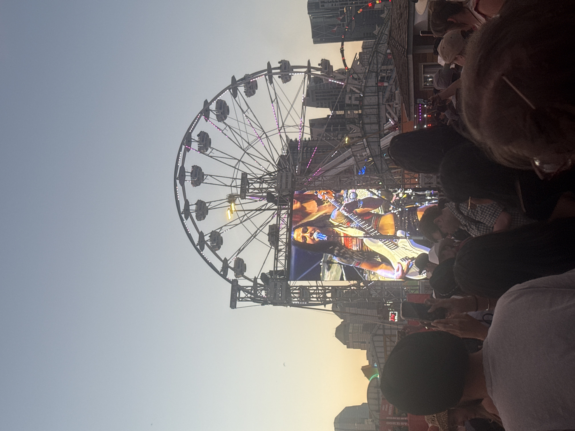The image depicts a nighttime outdoor concert at Stampede Park in Calgary, Canada. A large Ferris wheel with illuminated cabins is visible in the background, against a dusky sky. A giant screen displays a female musician playing a white electric guitar and singing into a microphone, dressed in a colorful outfit. In front of the screen, a crowd of people are gathered, many holding up their phones to record the performance. The foreground is filled with the backs of concert-goers. The overall scene suggests a lively and energetic atmosphere at a festival or major event.