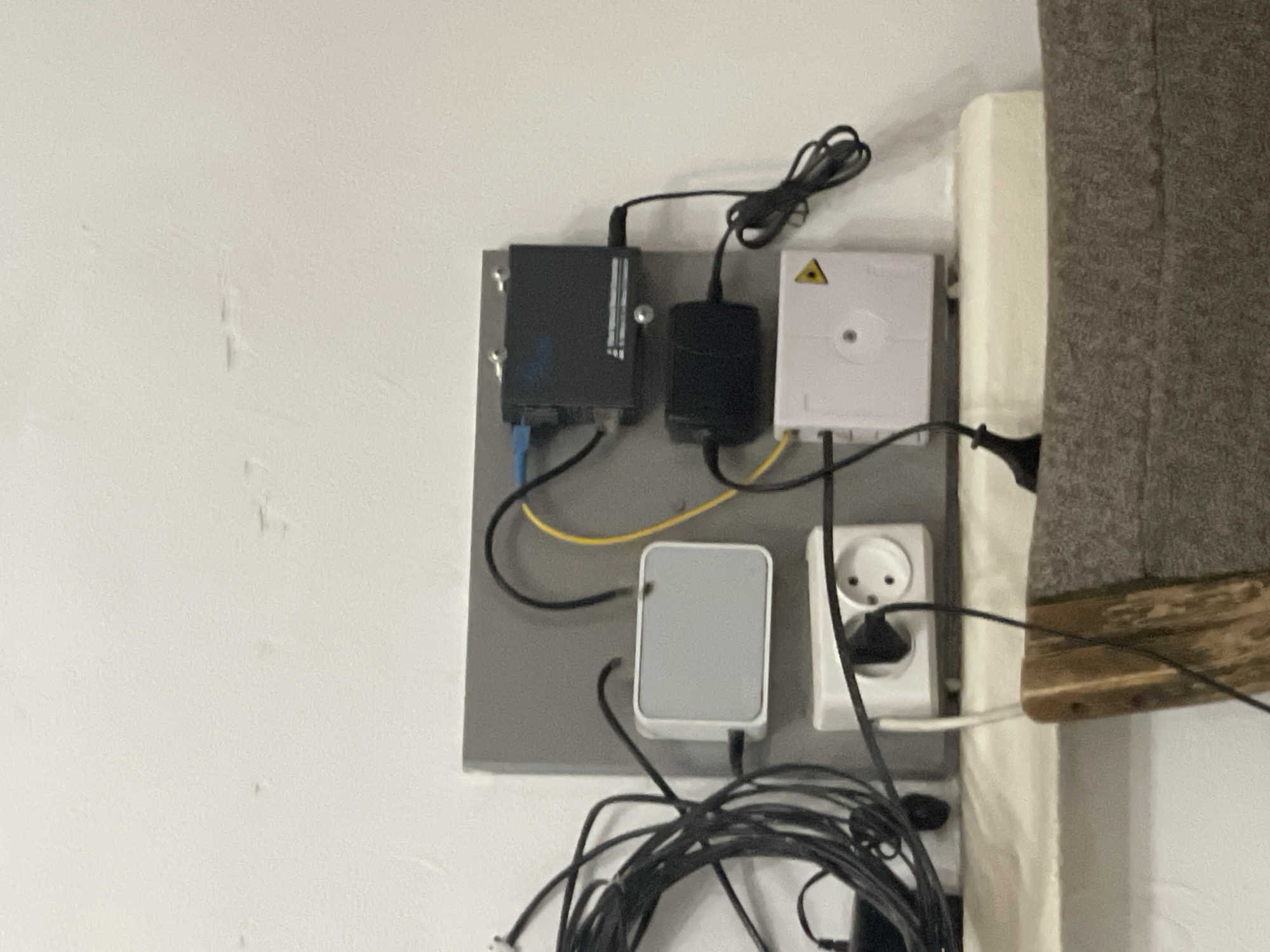 The image shows a wall-mounted setup of electronic devices and cables. There are several black cables bundled together on the left side of the image. Mounted on a grey board are a white rectangular device, a black rectangular device with a blue cable connected to it, another black rectangular device below it with a power cable, and a white rectangular device with a yellow triangle symbol on it. A white electrical outlet with a black plug inserted into it is mounted below the white rectangular device. The background is a plain, textured white wall. The scene is indoors, likely a residential or office space. There is no indication of the location being Bishkek City, Kyrgyzstan, or any specific city.
