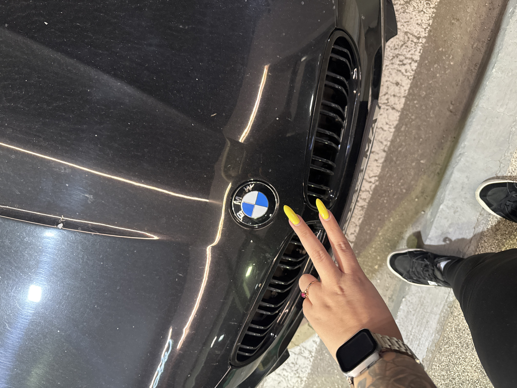 The scene depicts a partial view of a black BMW automobile, observed from an elevated perspective. The location is specified as the city of Krepost, Bulgaria.
**Vehicle:**
The front section of a black BMW is centrally positioned. The car features the standard BMW roundel emblem on the hood and a black kidney grille with vertical slats. The hood surface exhibits numerous fine dust particles and reflects strong, linear light sources, revealing faint surface imperfections or scratches. The bumper and lower grille area are also black.
**Person:**
A single individual is partially visible on the left side of the frame, positioned beside the vehicle.
- **Right Hand and Forearm:** A right hand is extended forward, displaying a "V" sign with the index and middle fingers. The fingernails are long, almond-shaped, and colored bright yellow. A gold-toned ring with a single red stone is present on the ring finger. The forearm displays dark, extensive tattoos. A silver-colored smartwatch with a metallic link band is worn on the wrist; its screen is dark.
- **Left Leg and Foot:** The individual's lower left leg is visible. They are wearing black trousers, black sneakers with white soles and laces, and white socks. The foot is positioned on a textured, light-colored ground surface.
**Environment:**
The ground consists of a mixed surface. Immediately below the car and extending to the right is a dark, textured surface, possibly asphalt or a coarse material. To the left, beneath the person's foot, is a lighter, finely textured concrete or paved surface, separated by a distinct linear curb. The ambient lighting appears dim, with bright, elongated reflections on the car's surface, indicating artificial light sources above or in front of the vehicle.
