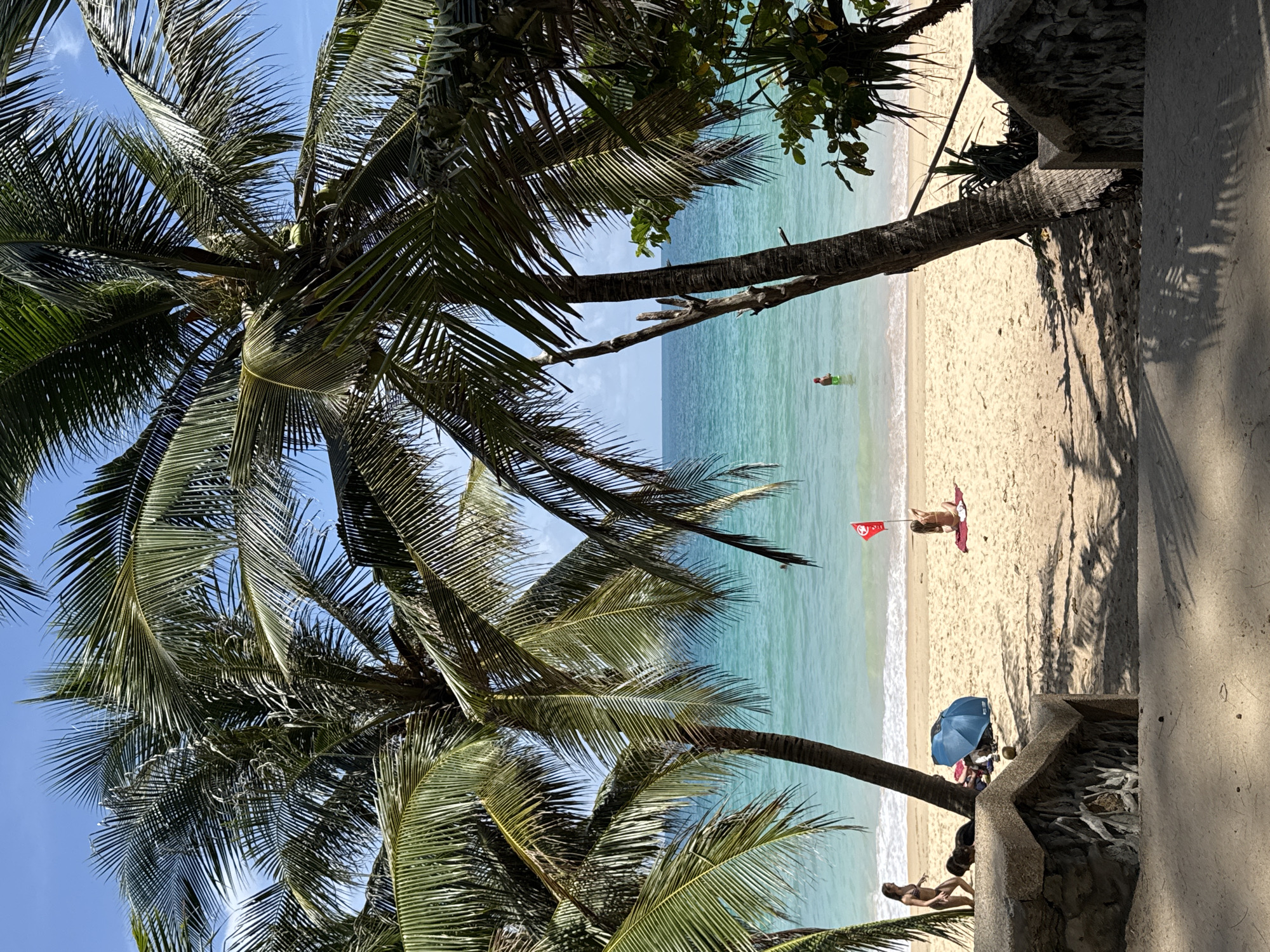 This is a daytime, eye-level shot of a tropical beach in Rawai, Thailand, viewed through the fronds of palm trees. The foreground features a stone staircase with rough-hewn stone walls leading down towards the sand. Sunlight casts distinct shadows of palm fronds onto the sand and the steps. The beach itself is light beige sand with gentle waves lapping at the shore.
Several people are visible on the beach. On the left, two women are walking towards the water; one is wearing a bikini and the other is walking behind her. Further down the beach, near a blue umbrella, at least two other people are seated or reclining. To the right of the umbrella, a person in a bikini is sitting on a red towel, holding a red flag on a stick aloft. In the turquoise ocean, further out, a person wearing a green hat or swim cap is wading.
The ocean is a clear turquoise color, transitioning to a deeper blue towards the horizon. The sky is a bright, clear blue with a few wispy white clouds. The palm trees in the foreground are lush and green, their fronds creating a natural frame for the beach scene.