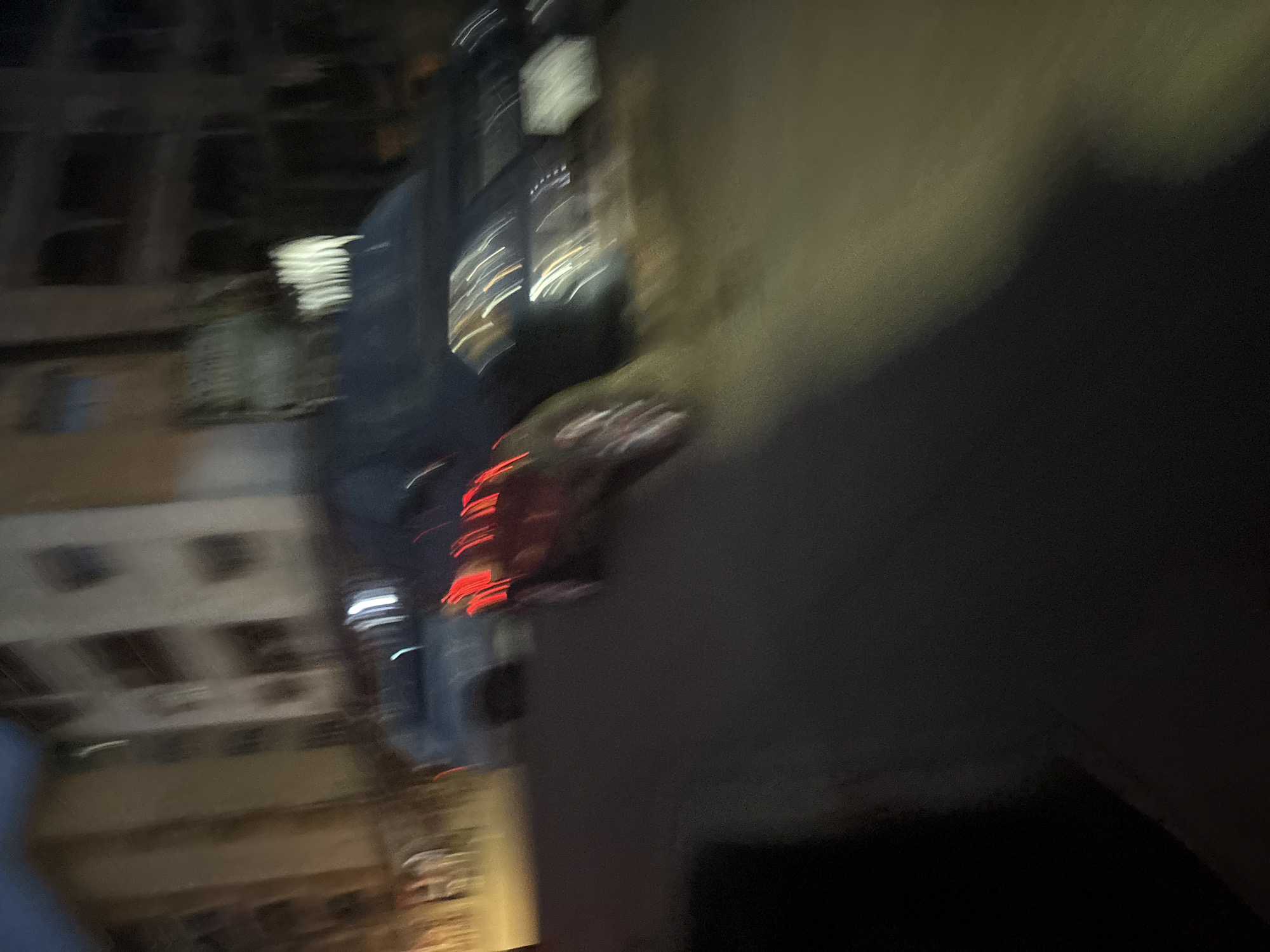 The image captures a blurry nighttime street scene in Onitsha, Nigeria, with a building on the left and a road in the foreground. Two vehicles are visible on the road: a dark SUV in the middle, partially covered by what appears to be a tarp or plastic sheeting, and a red vehicle to its left, with its brake lights illuminated, indicating it may be stationary or moving slowly. The red vehicle's wheels are blurred, suggesting motion. The building on the left is multi-story with visible windows and balconies, appearing to be an apartment complex or commercial structure. The road surface is dark, and streetlights or vehicle headlights create streaks of light due to the motion blur. The overall impression is one of a busy urban environment at night, with limited clarity due to the low light conditions and camera movement.