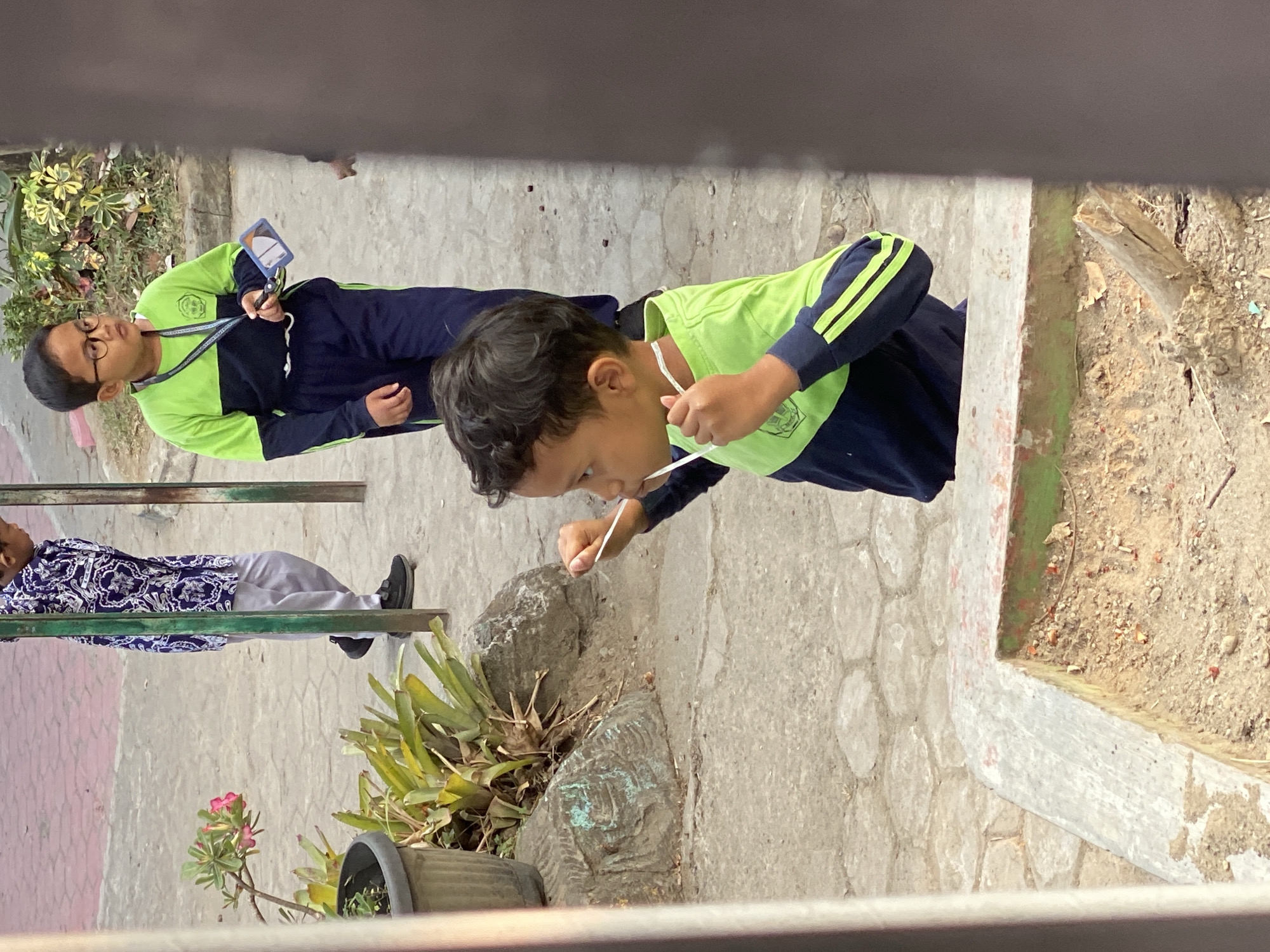 The image shows a close-up view of a young boy in the foreground, looking to his right and holding a white string in his hands, which appears to be a lanyard. He is wearing a long-sleeved shirt with a green top and navy blue sleeves, and navy blue pants. The background shows a paved area with hexagonal stones, a planter with greenery and a pink flower, and a large rock formation. In the middle ground, another boy wearing glasses and a similar uniform is visible, holding a card. Further in the background, a person in a blue and white patterned shirt and grey pants is walking away. The scene suggests an outdoor setting, possibly a schoolyard in Kasihan, Indonesia, given the context.