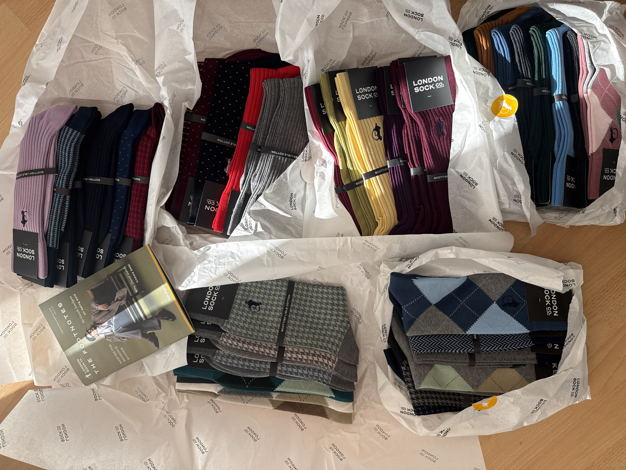 The scene shows five separate bundles of socks, each wrapped in white tissue paper imprinted with the "London Sock Co." logo. Each bundle contains multiple pairs of socks, visible in a variety of colors and patterns. The socks are held together with a black paper band printed with the company's name. A book titled "The Footnotes" is placed near one of the bundles. The background consists of a light-colored wooden surface. There is no information about the location or people in the city of London.