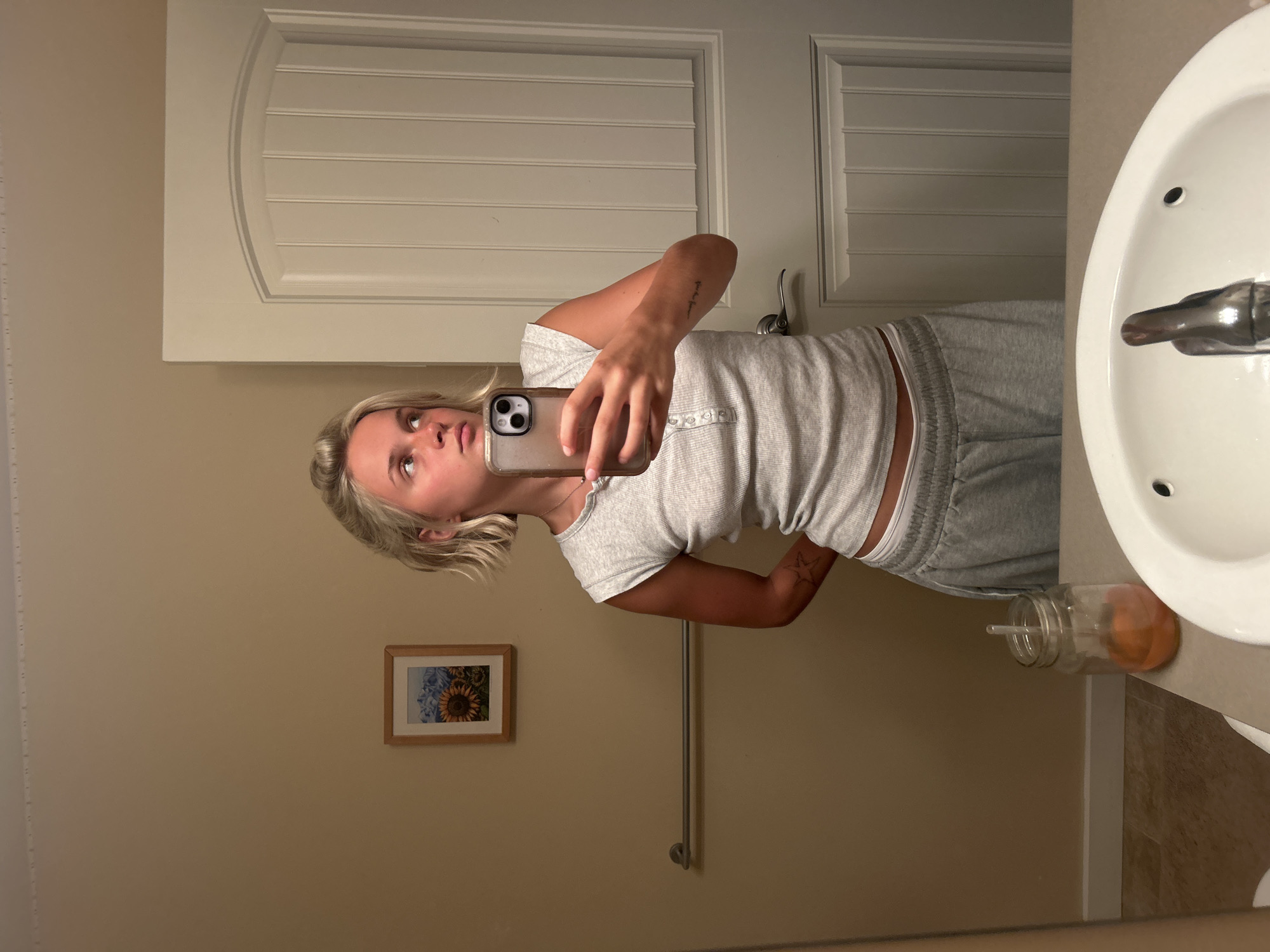 A young woman with blonde hair, wearing a grey tank top and grey sweatpants, stands in a bathroom and takes a mirror selfie. She is looking to the right, away from the camera. Behind her, a closed white door is visible. To her left, a small framed picture of sunflowers and mountains hangs on a tan wall, with a towel rack below it. A glass jar with an orange liquid and a straw sits on the counter in front of the sink.