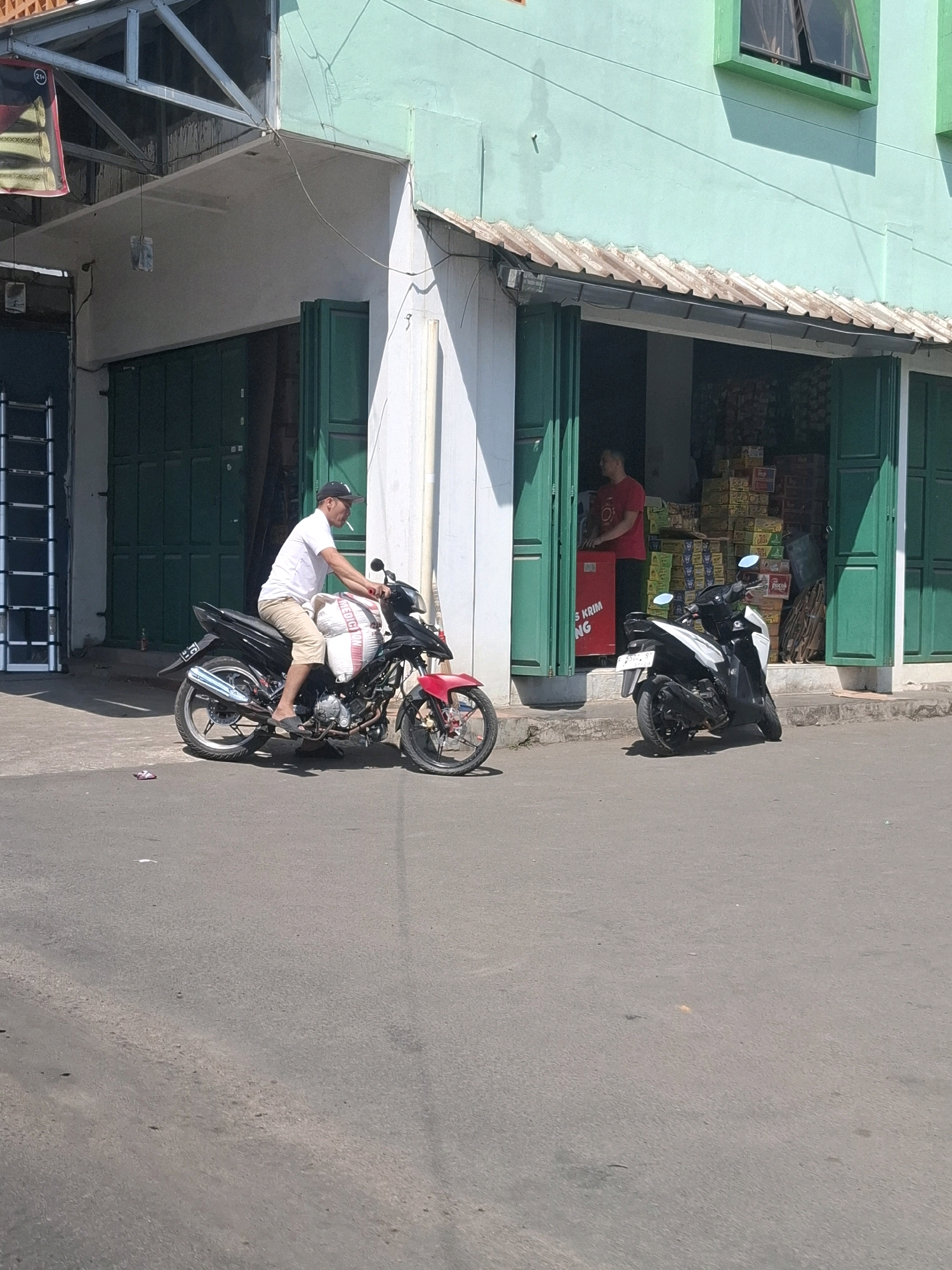A man wearing a white t-shirt, tan shorts, a black baseball cap, and black flip-flops is seated on a black motorcycle. The motorcycle has a large, white sack secured to its rear. The man is holding the handlebars, and a cigarette is visible in his mouth. The motorcycle is parked on a paved street in front of a storefront. The storefront features closed green metal doors and an awning. Inside the store, shelves are stocked with boxes, and another person wearing a red shirt is visible. A second, white and black motorcycle is parked in front of the store. The scene takes place outdoors under a bright sky, with shadows cast by the buildings. The location is identified as Gunung Guruh, Indonesia.