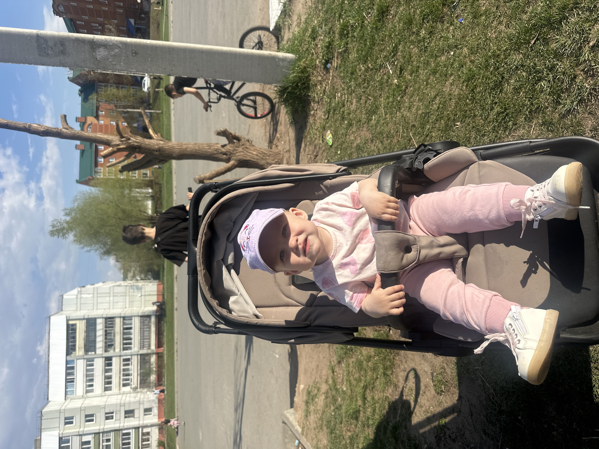 A toddler wearing a purple cap, pink leggings, a white shirt with pink floral patterns, and white shoes sits in a stroller outside in Chaikovsky, Russia. A person rides a bicycle in the background, and there is a building complex in the background. There is a utility pole and a bare tree to the left of the bicycle. Grass covers a small patch of land to the right of the stroller. The image shows the child looking toward the camera and smiling.