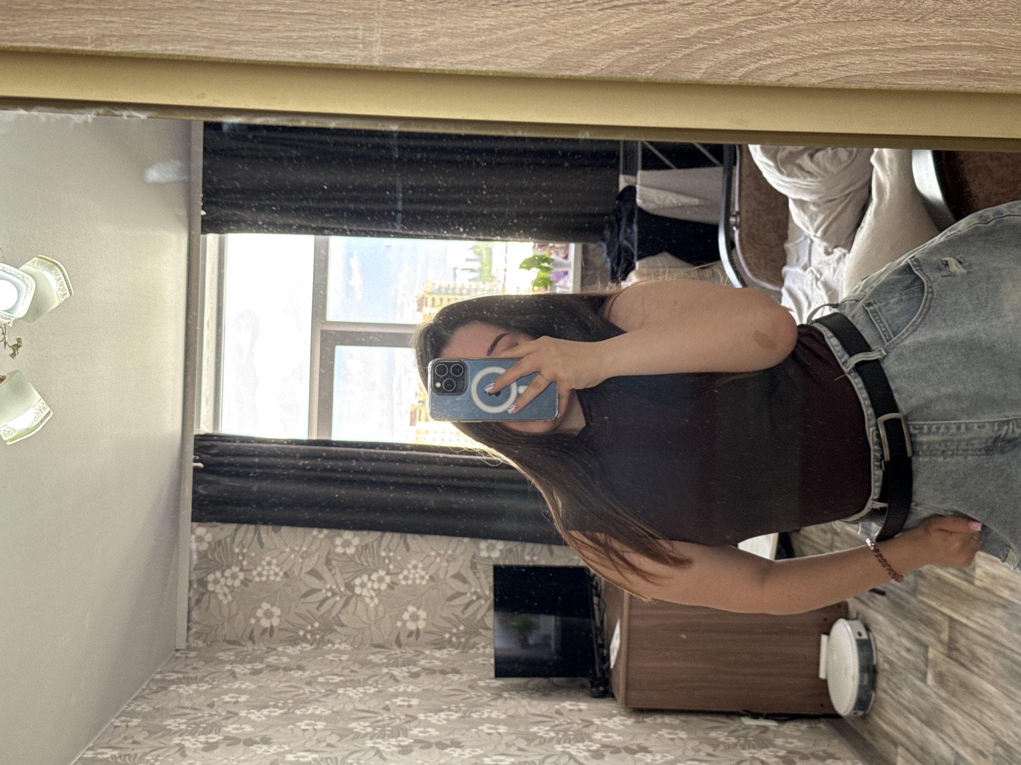 The image is a selfie taken in a bedroom. A young woman is reflected in a large mirror, holding a smartphone to take the picture. She is wearing a dark sleeveless top, a black belt, and light-wash denim shorts with some distressing. A beaded bracelet is visible on her left wrist. In the background, there is a window with dark curtains, a wooden dresser with a TV on it, and a robot vacuum cleaner on the floor. The room has patterned wallpaper and a wooden floor. The scene appears to be in Zarechnaya (Gamovskoe S/P), Russia.
