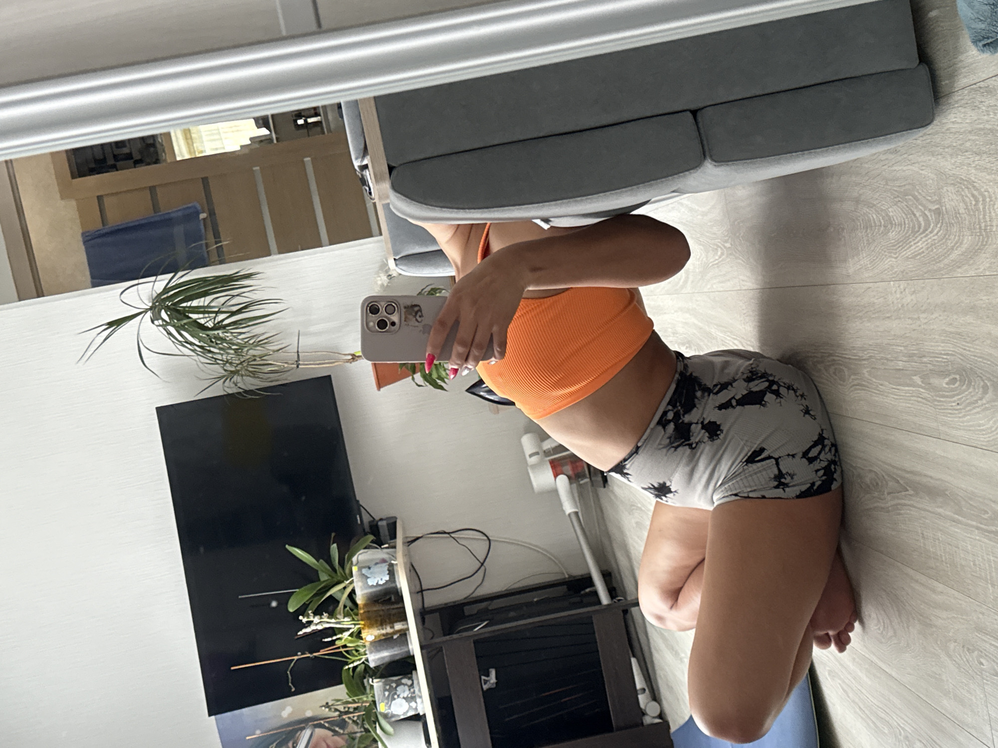 A woman is taking a selfie in a mirror. She is wearing an orange ribbed tank top and gray and black tie-dye shorts. The reflection shows a gray sofa, a TV, a plant, and a TV stand. A vacuum cleaner is visible near the stand. The floor is light-colored wood or laminate. There is no information about the location being in Kauchuk Stantsiya, Russia.