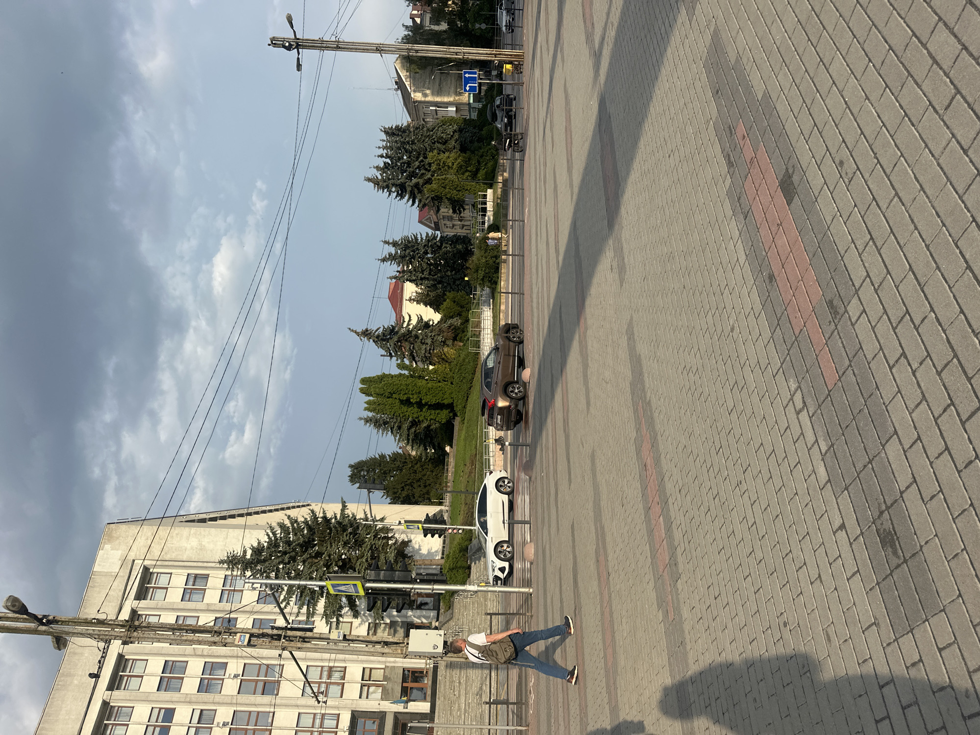 The image depicts a street scene in Ternopil, Ukraine, on a cloudy day. In the foreground, a man with a backpack is walking across a pedestrian crossing paved with grey and red bricks. To his left, a white Audi e-tron is stopped at a traffic light. Behind the pedestrian crossing, several cars are visible, including a brown SUV. The background features a large, light-colored building with many windows, flanked by tall evergreen trees. A traffic light and a street lamp are also present. The sky is overcast with patches of blue visible. The scene captures an everyday moment in the city, with a focus on the urban infrastructure and a few vehicles and a pedestrian.