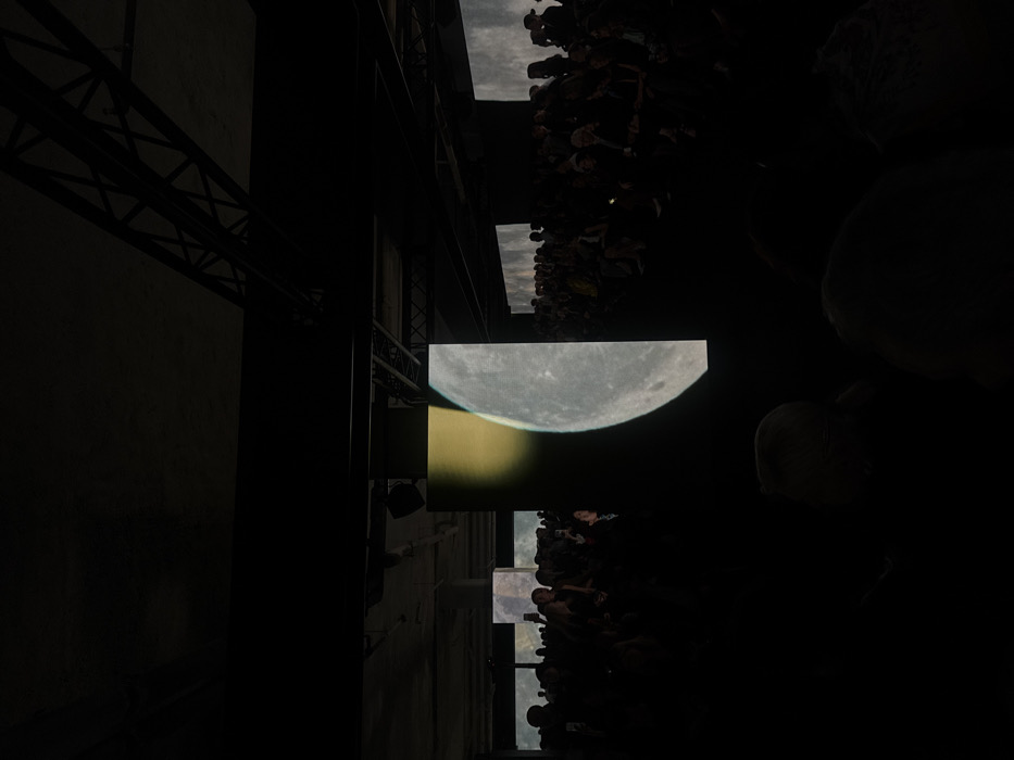 The image captures an indoor event held in a large, dimly lit, industrial-style hall, consistent with renovated exhibition or event spaces found in Paris, France. The high ceiling features exposed dark metal trusses and concrete beams. A substantial audience is present, with numerous individuals seated in rows, their forms largely visible as dark silhouettes against the ambient light from digital displays. Their collective attention is directed towards several large, vertical LED screens or projections. The most prominent screen, centrally positioned, displays a detailed image of a large celestial body; it is grey, cratered, and illuminated on its right side, with a distinct yellowish-orange crescent partially visible behind its upper left quadrant. Additional screens in the background show similar celestial or abstract landscape imagery. Several audience members are holding up smartphones, indicating they are recording or photographing the event. The environment is dark and immersive, dominated by the illuminated digital projections.